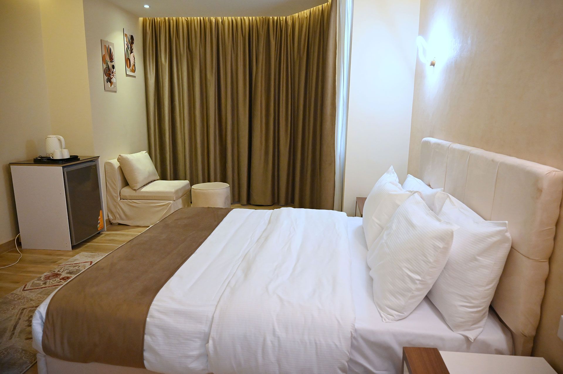 Bedroom interior with a bed, small fridge, and sitting area. Cream and brown tones.