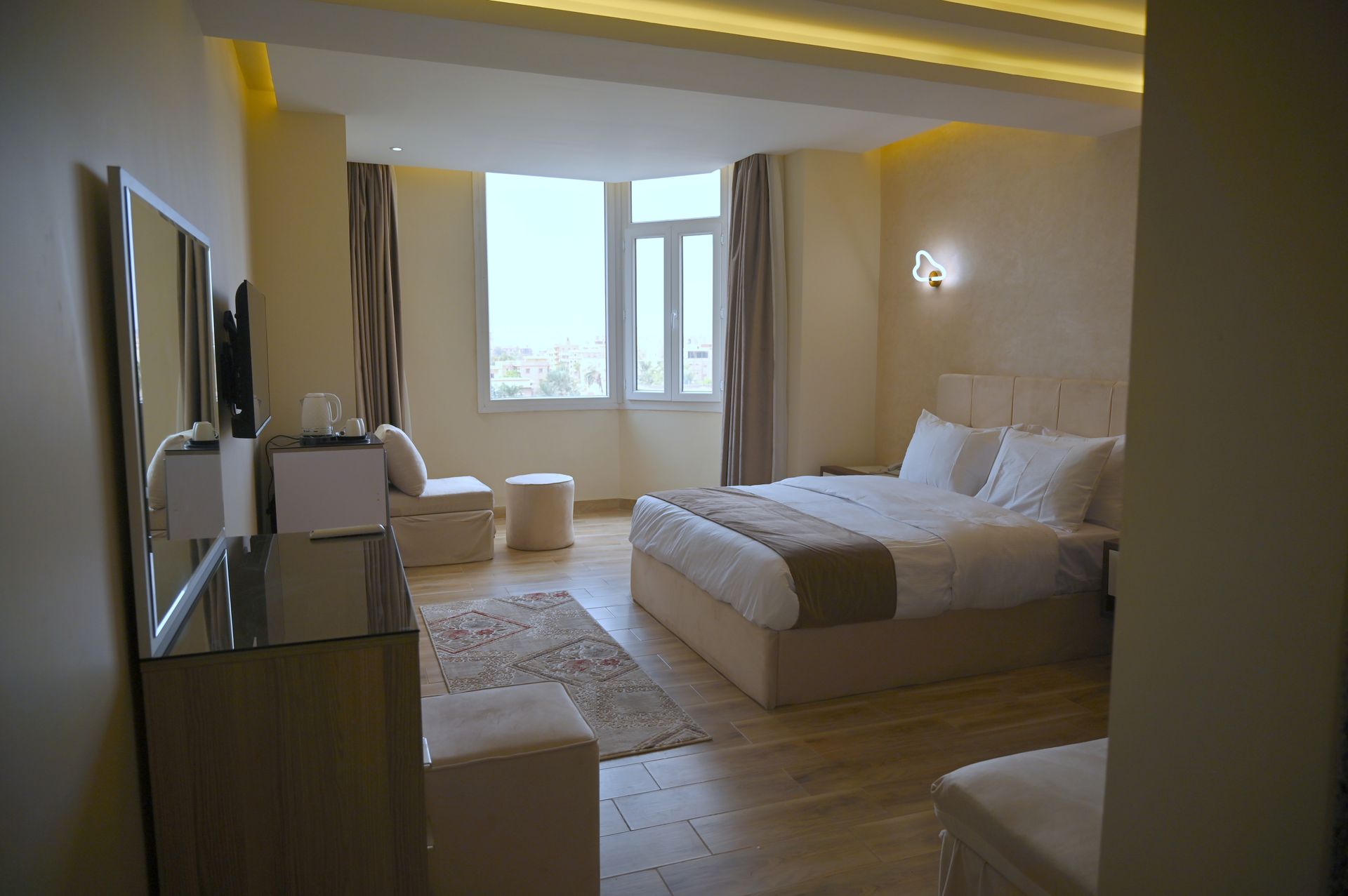 Hotel bedroom with bed, window, seating, and dresser. Light walls and flooring.