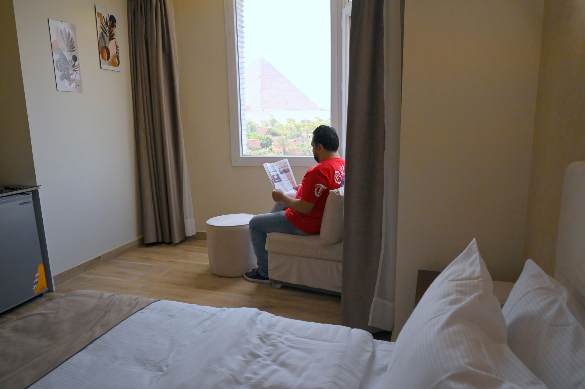 Man sitting in a chair by a window, reading a paper. A bed and mini-fridge are in the room.