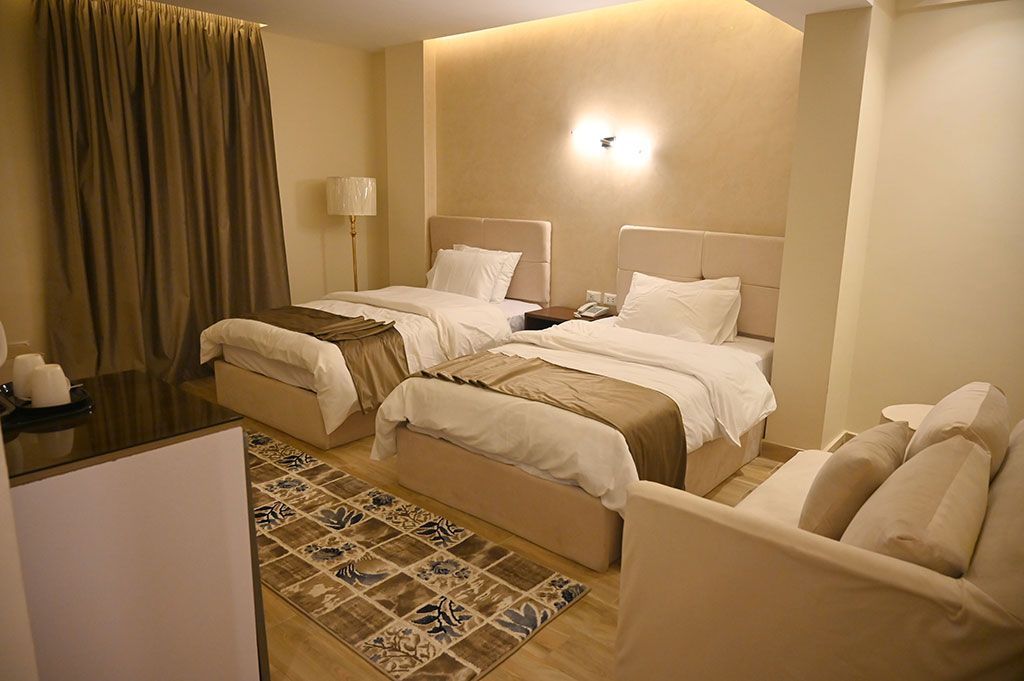 Hotel room with two twin beds, small sofa, and patterned rug. Warm neutral tones.