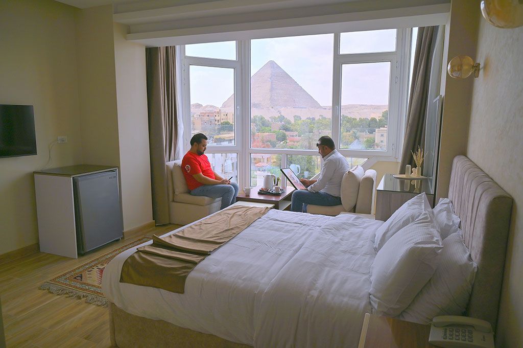 Hotel room with pyramid view; two people sit by window, bed in foreground.