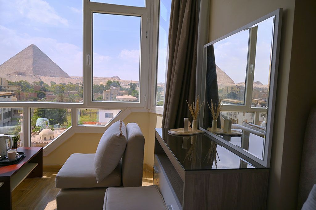 Room with pyramid view: window, seating, vanity with mirror, and the Great Pyramid in the background.