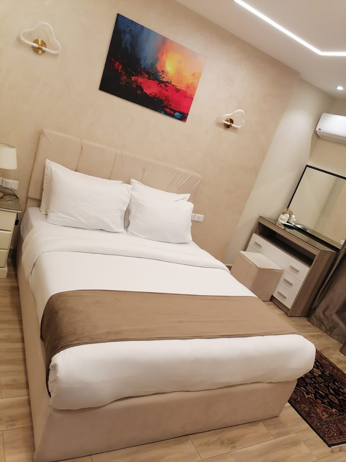 Bedroom with bed, side table, mirror, and decorative items. Soft neutral tones and wood accents.