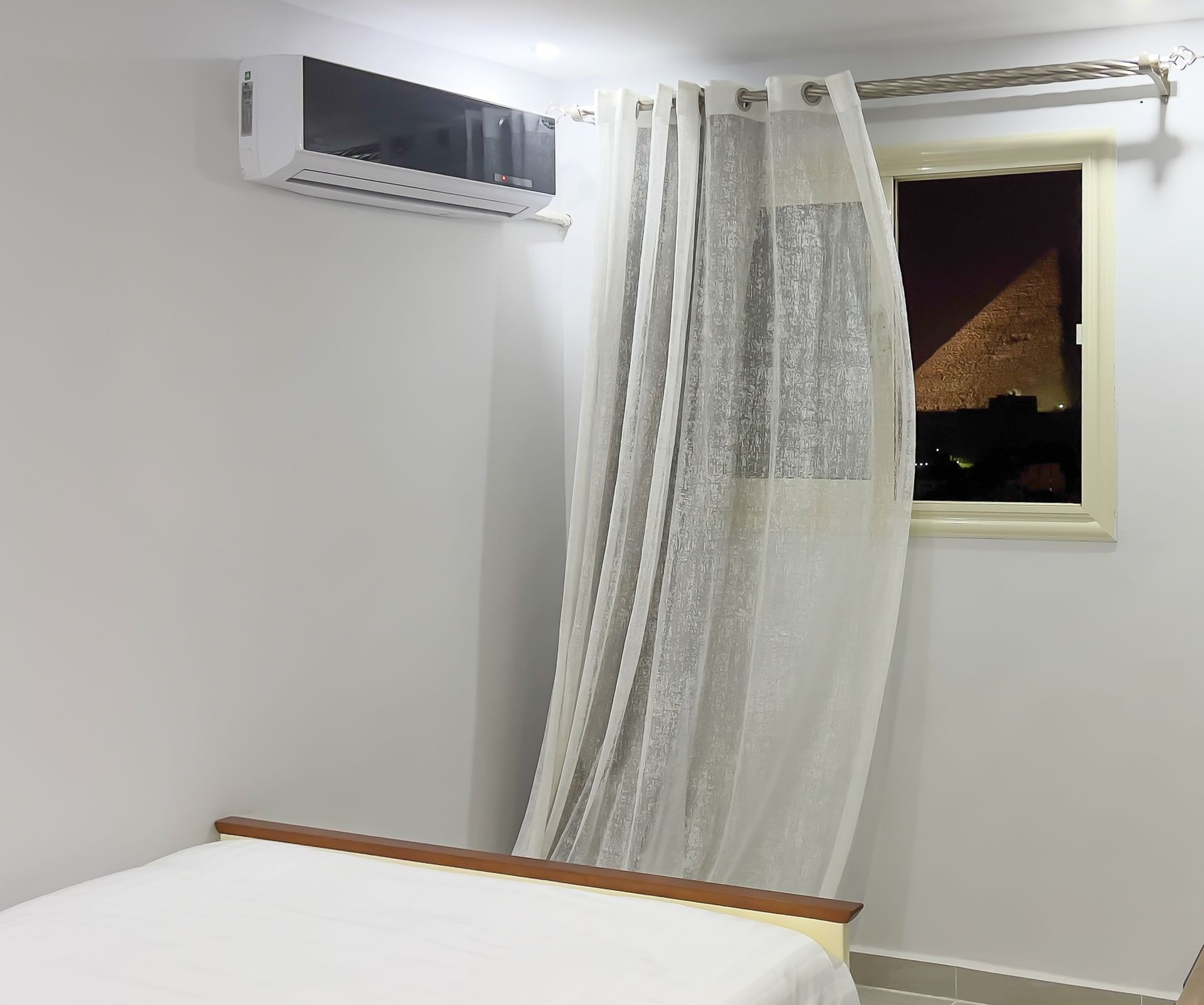 A bedroom with an air conditioner, a window with a sheer curtain, and the pyramids visible at night.