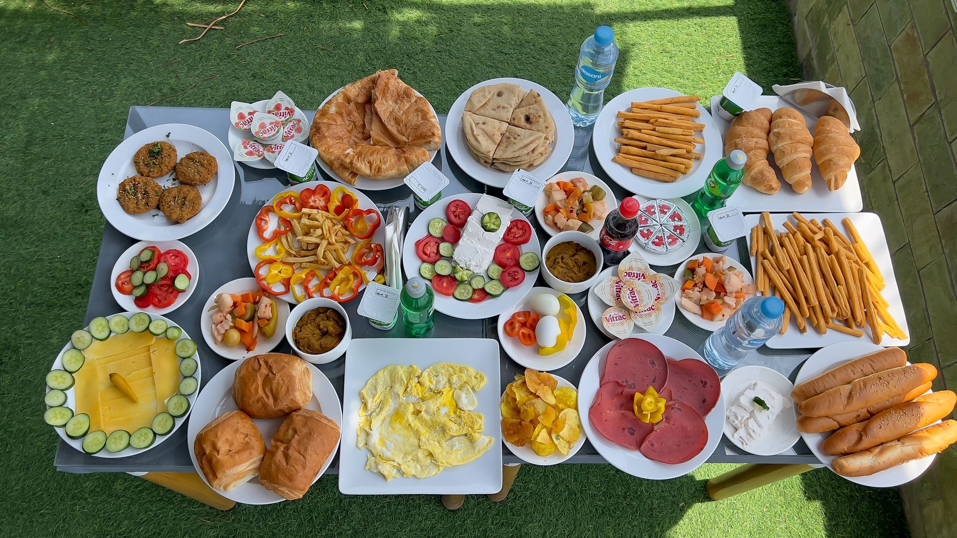 Table set with diverse food items: bread, eggs, vegetables, pastries, and drinks. Outdoors on green surface.