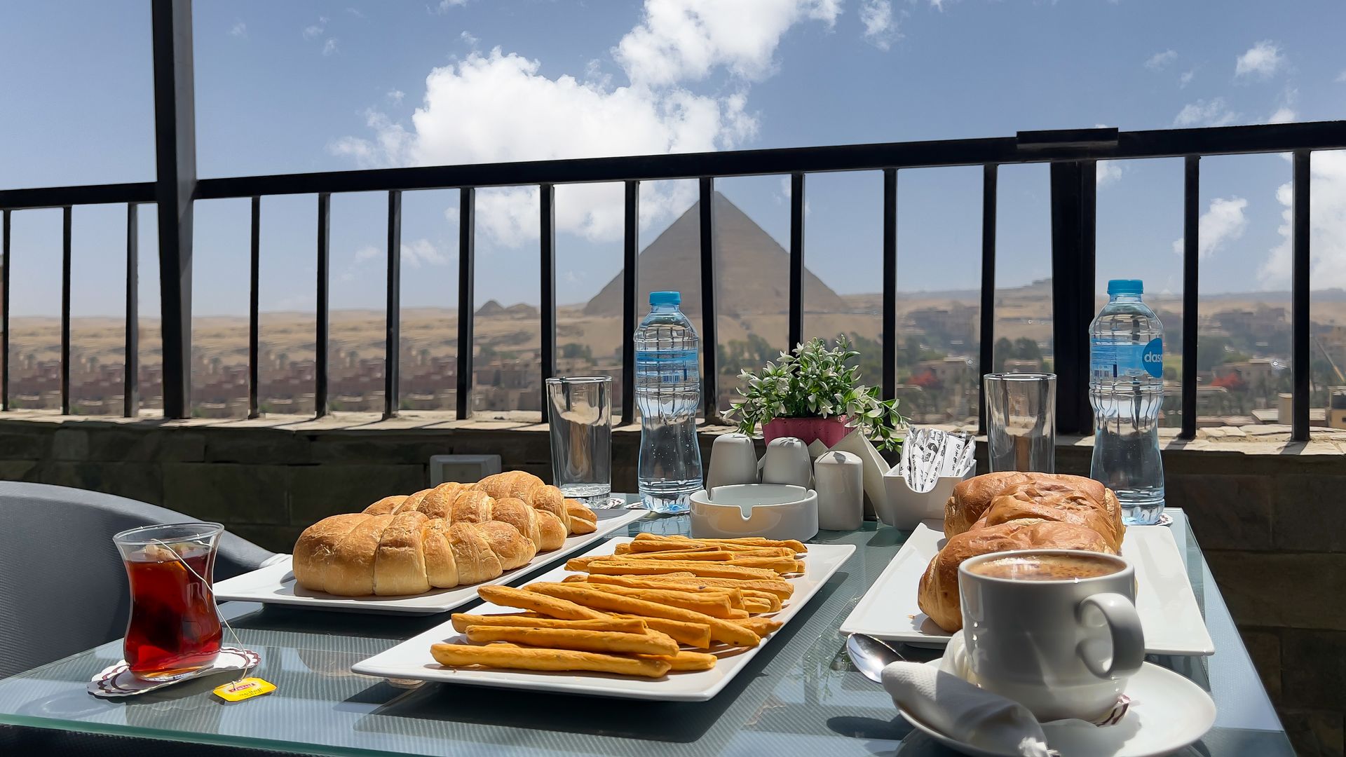 Breakfast spread with pyramids in background. Bread, fries, coffee, and water bottles on a table with a balcony view.