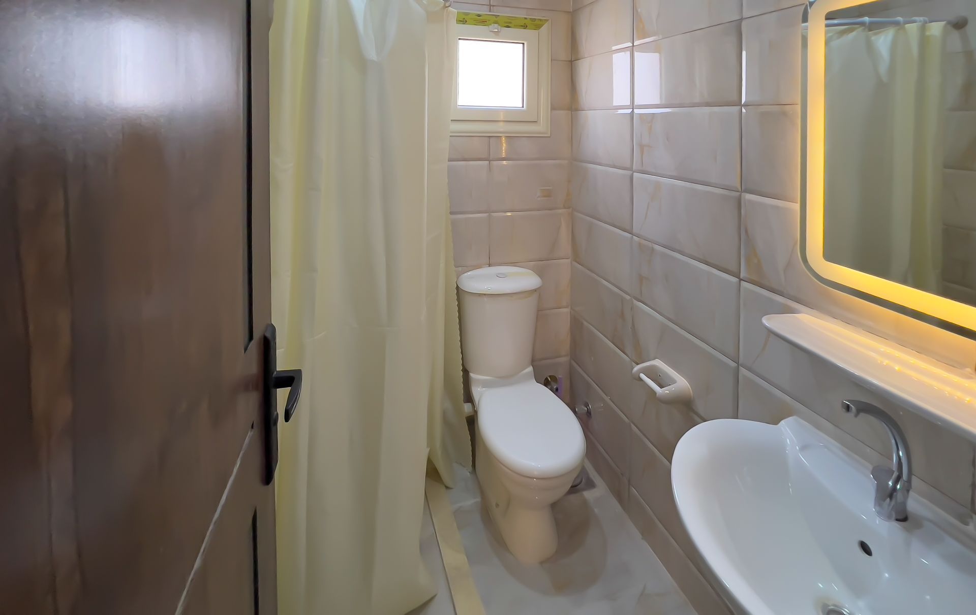 Bathroom with toilet, sink, and shower curtain. Light beige tiled walls, small window, and a door.