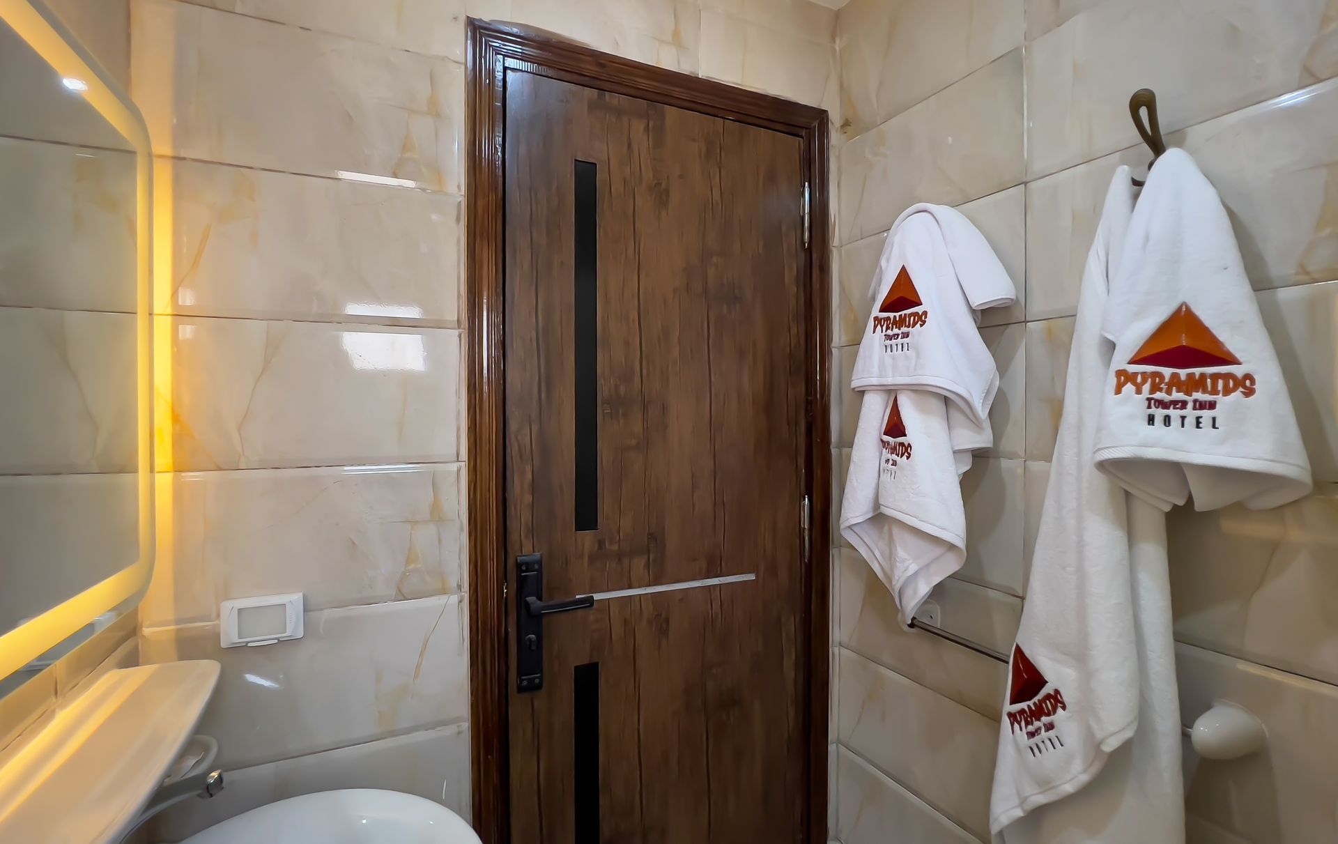Bathroom with brown door, white towels with pyramid logo, beige tiled walls, and a sink.
