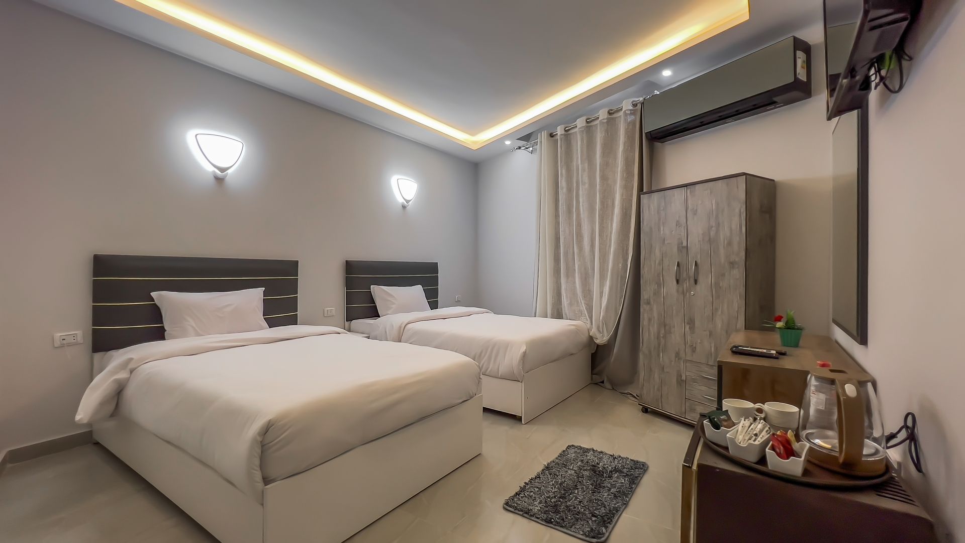 Twin-bed hotel room with neutral decor, two beds, a cabinet, and a side table with refreshments.