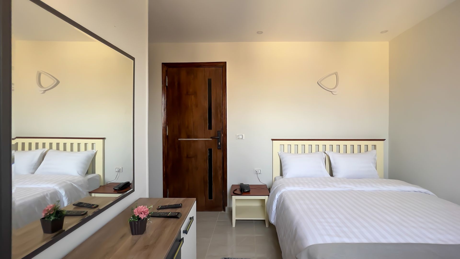 Bedroom with double bed, large mirror, wooden door, and nightstand. Light-colored walls and bedding.