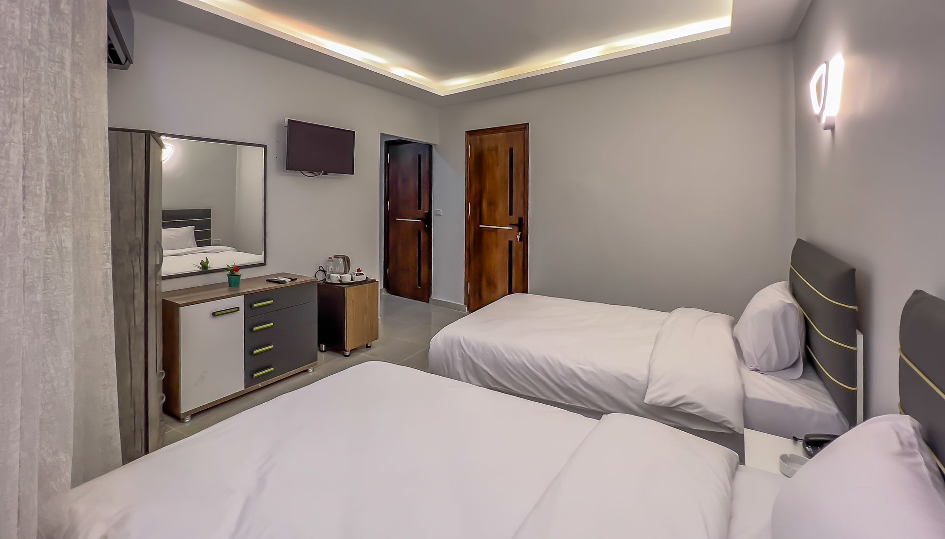 Two beds in a modern hotel room, with a dresser, mirror, and two closed wooden doors.