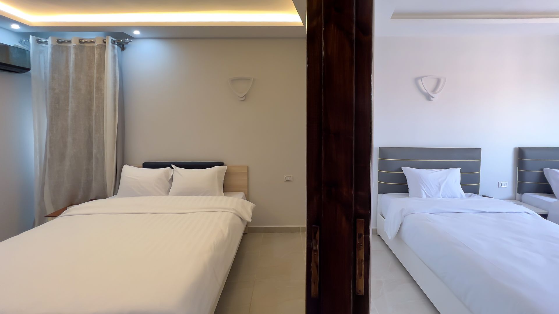 Two hotel bedrooms, one with a bed partly visible behind a wooden divider; both have white bedding, headboards, and walls.
