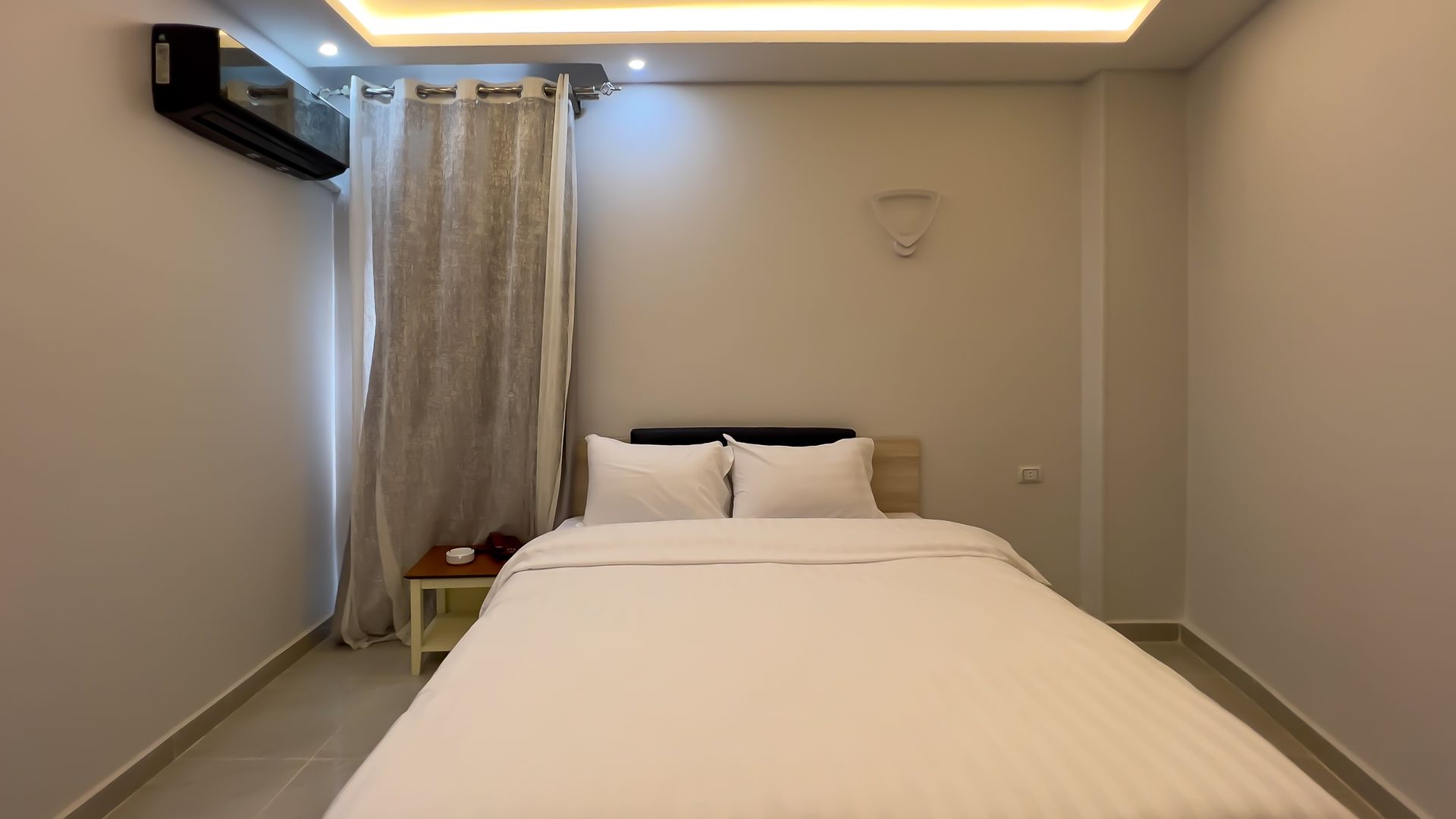 Bedroom with a double bed, white bedding, beige walls, and a window with curtains.