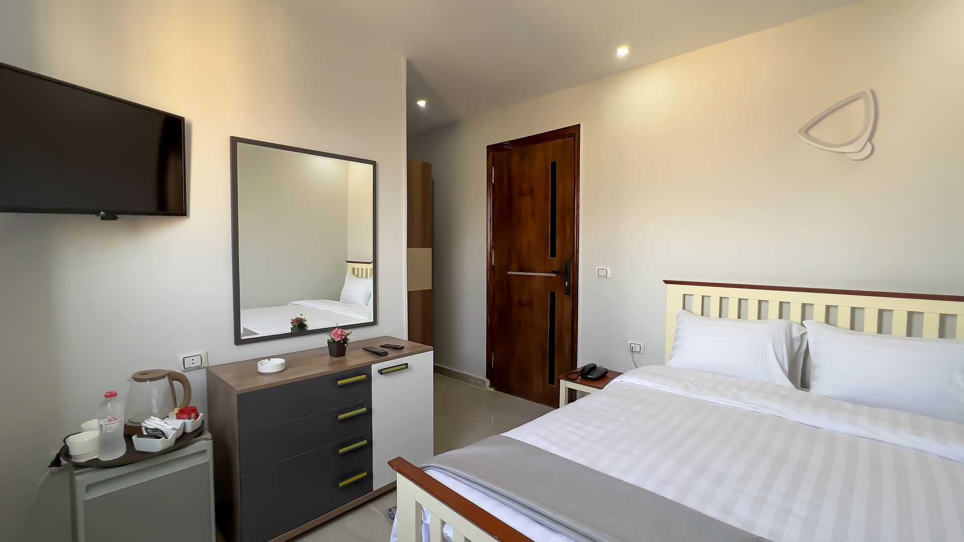 Hotel room with bed, dresser, mirror, TV, and closed wooden door. Neutral colors.