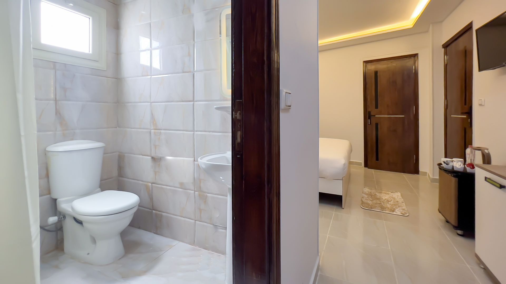 Bathroom doorway opening into a bedroom. Toilet on left. Bed and door in the room. Bright, clean space.