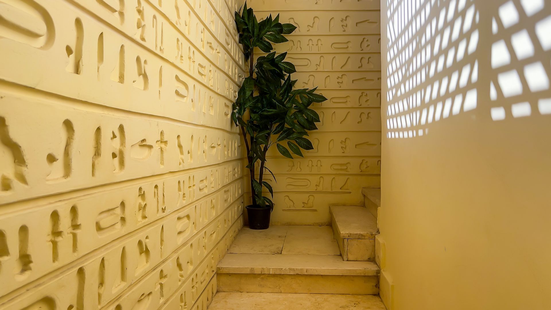 Stairwell with hieroglyphic walls, a potted plant, and light from a patterned window.