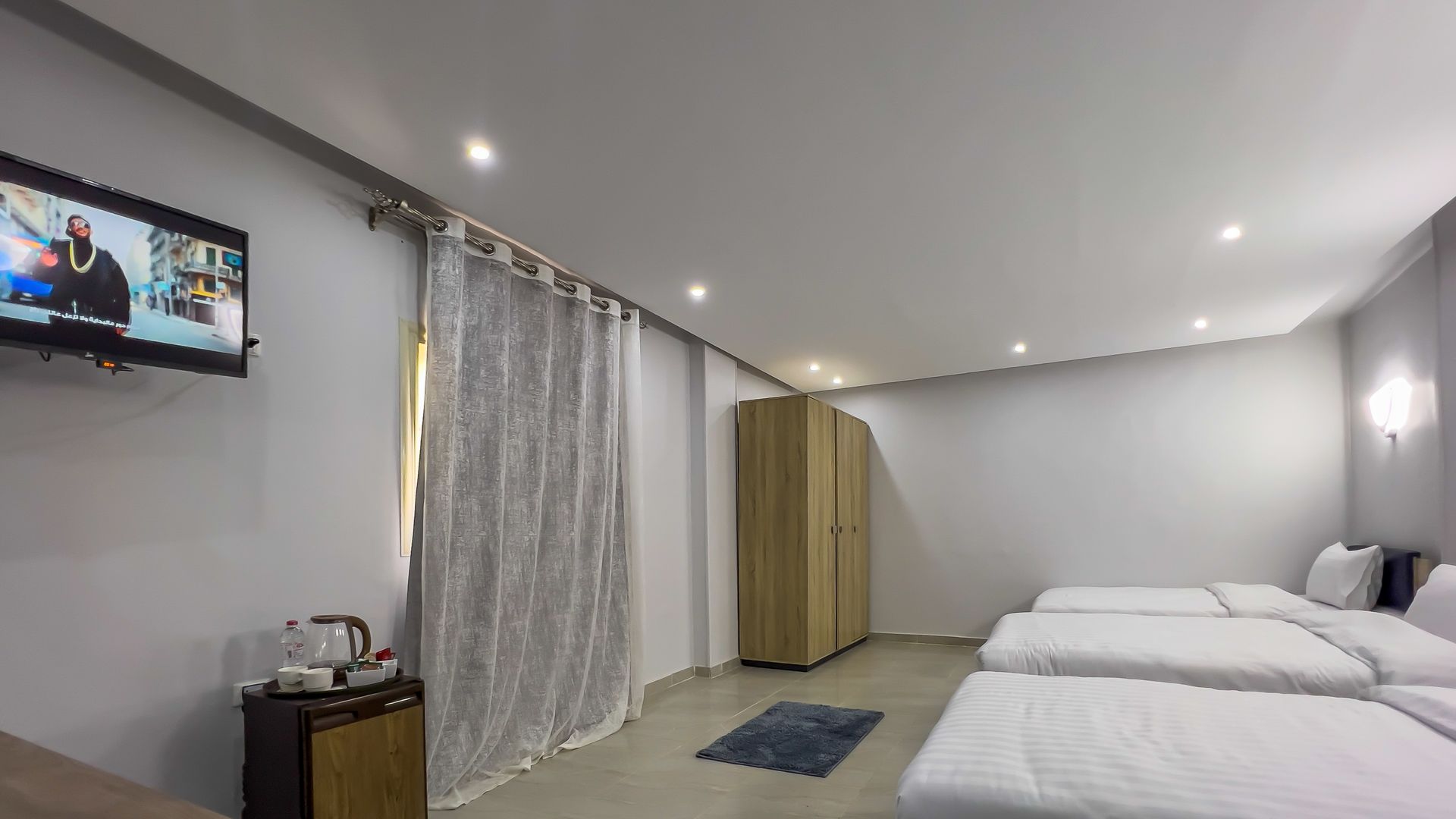 Hotel room with three beds, TV, wardrobe, and a small table with a kettle and tea set.