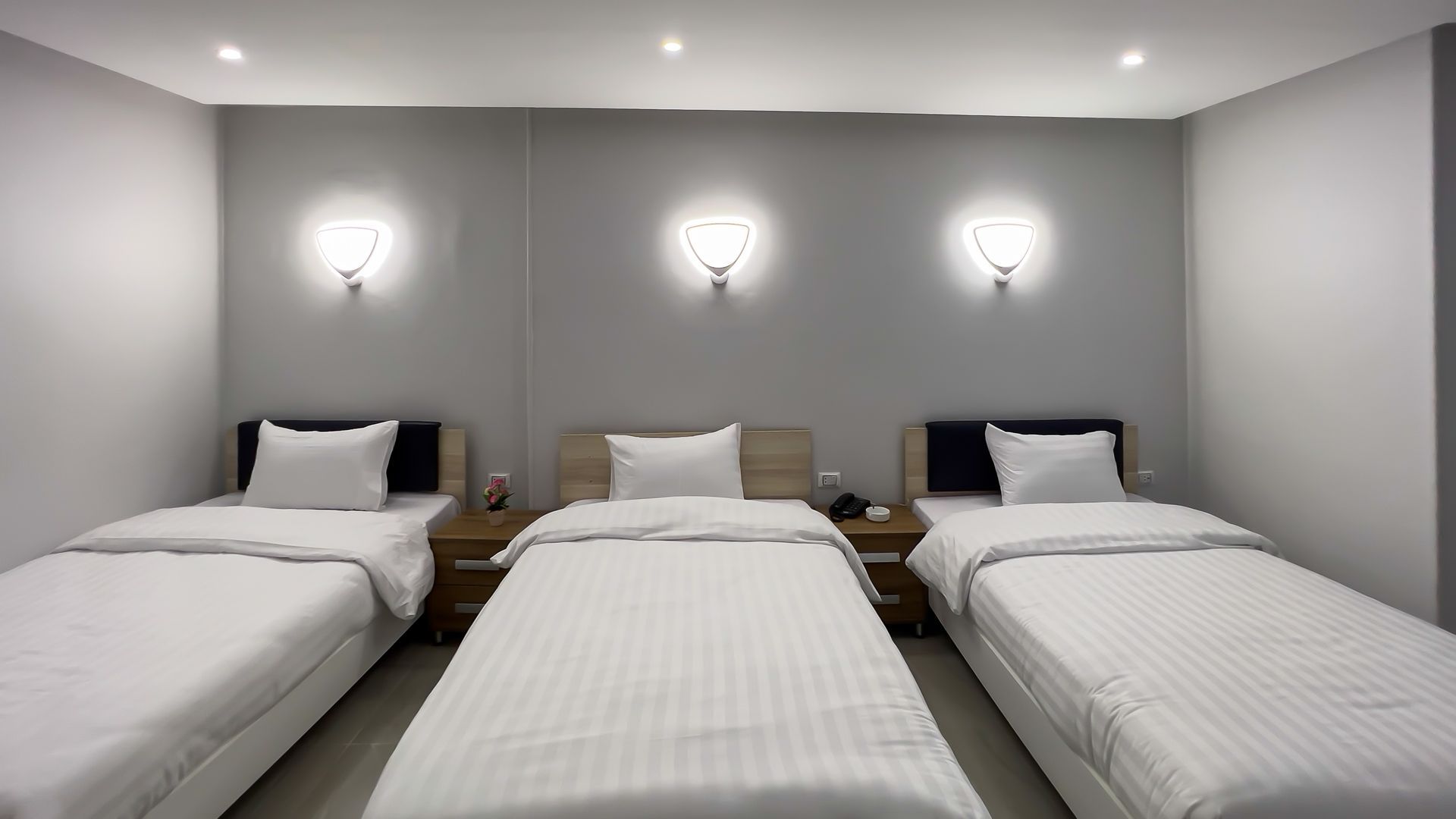 Three beds in a gray-walled room, each with white bedding. Wall sconces provide light.