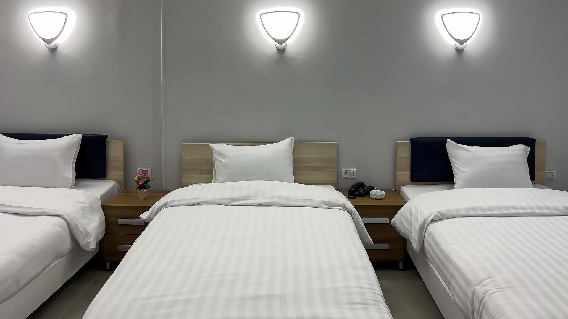 Three beds with white bedding against a gray wall, each with a bedside table and wall sconce.