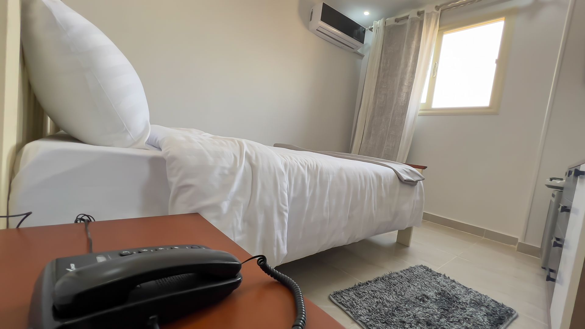 Bedroom with a single bed, white bedding, nightstand, phone, window, and air conditioner.