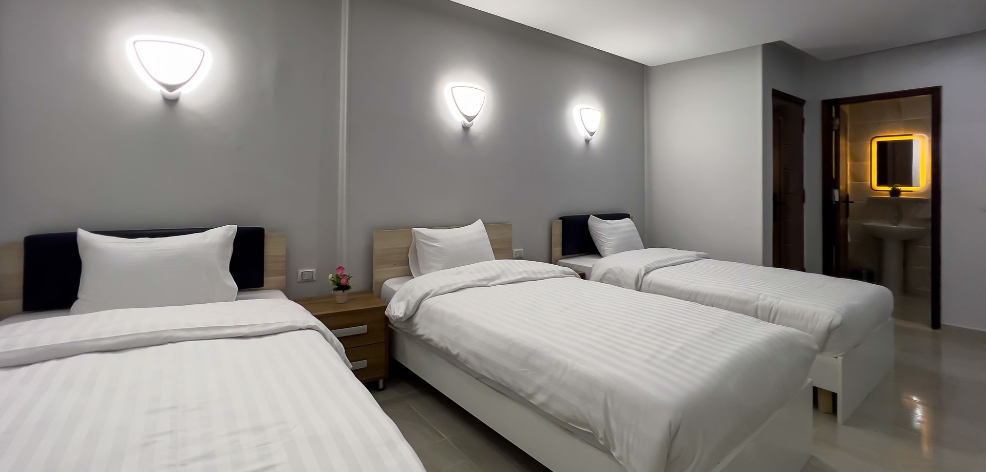 A hotel room with three single beds, grey walls, and a lit-up bathroom doorway.