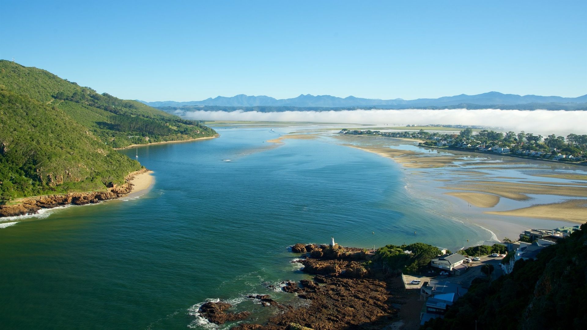 Blue waterway meets the ocean; lush green hills and shoreline on either side, distant mountains with low fog.