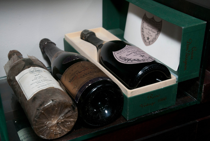 Three wine bottles, two appear aged. One is in a green box with the logo, one uncorked.
