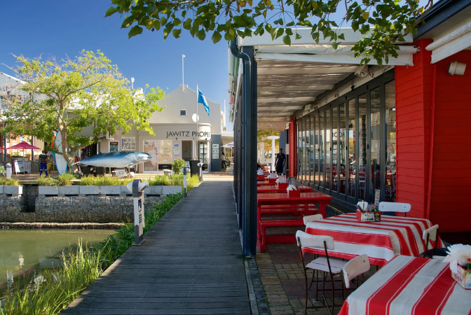Outdoor restaurant with red and white striped tablecloths, wooden walkway, and view of buildings and water.