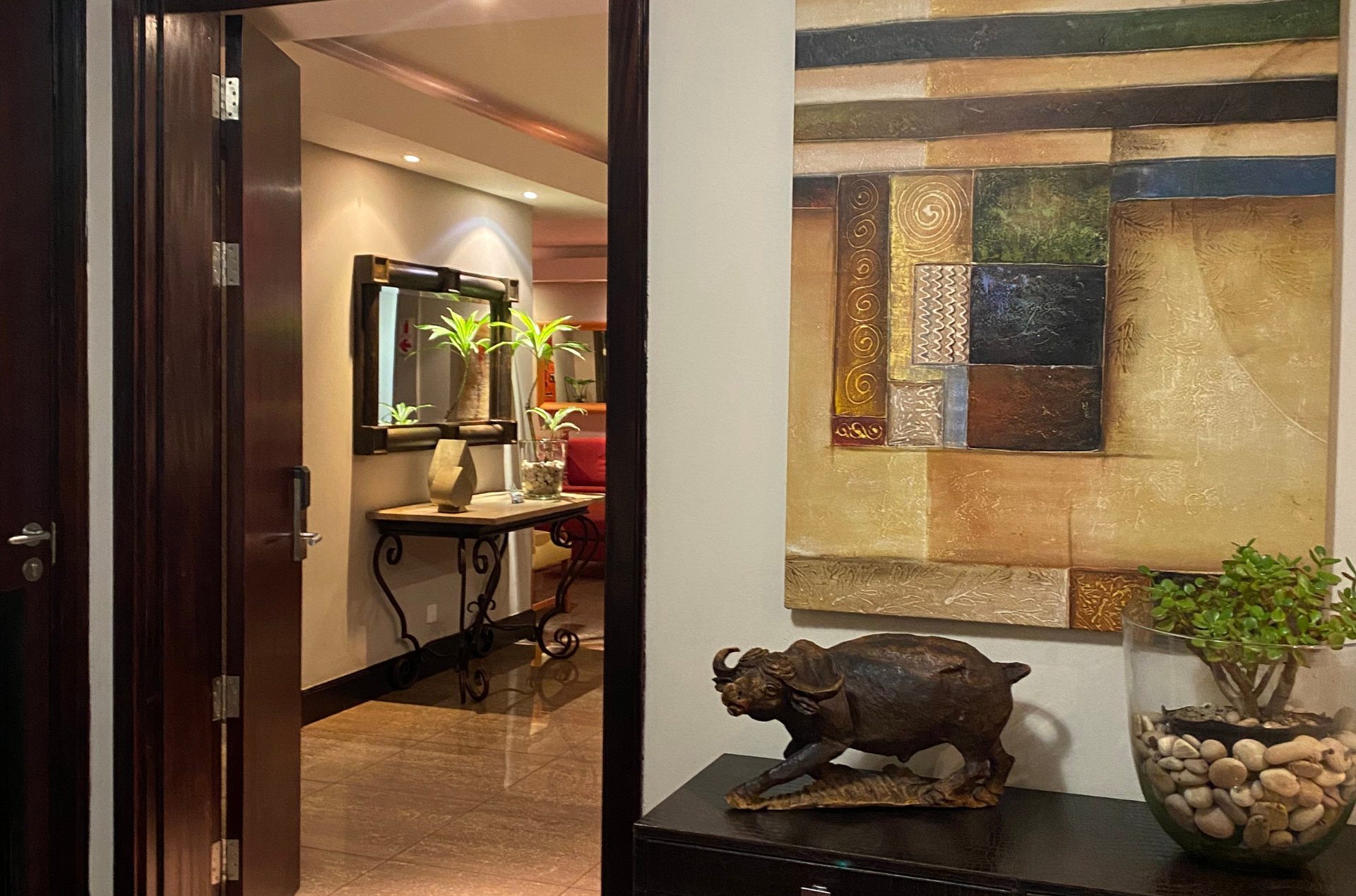 Open doorway reveals hallway with table, mirror, and artwork on a wall. Brown door and trim, marble floors.