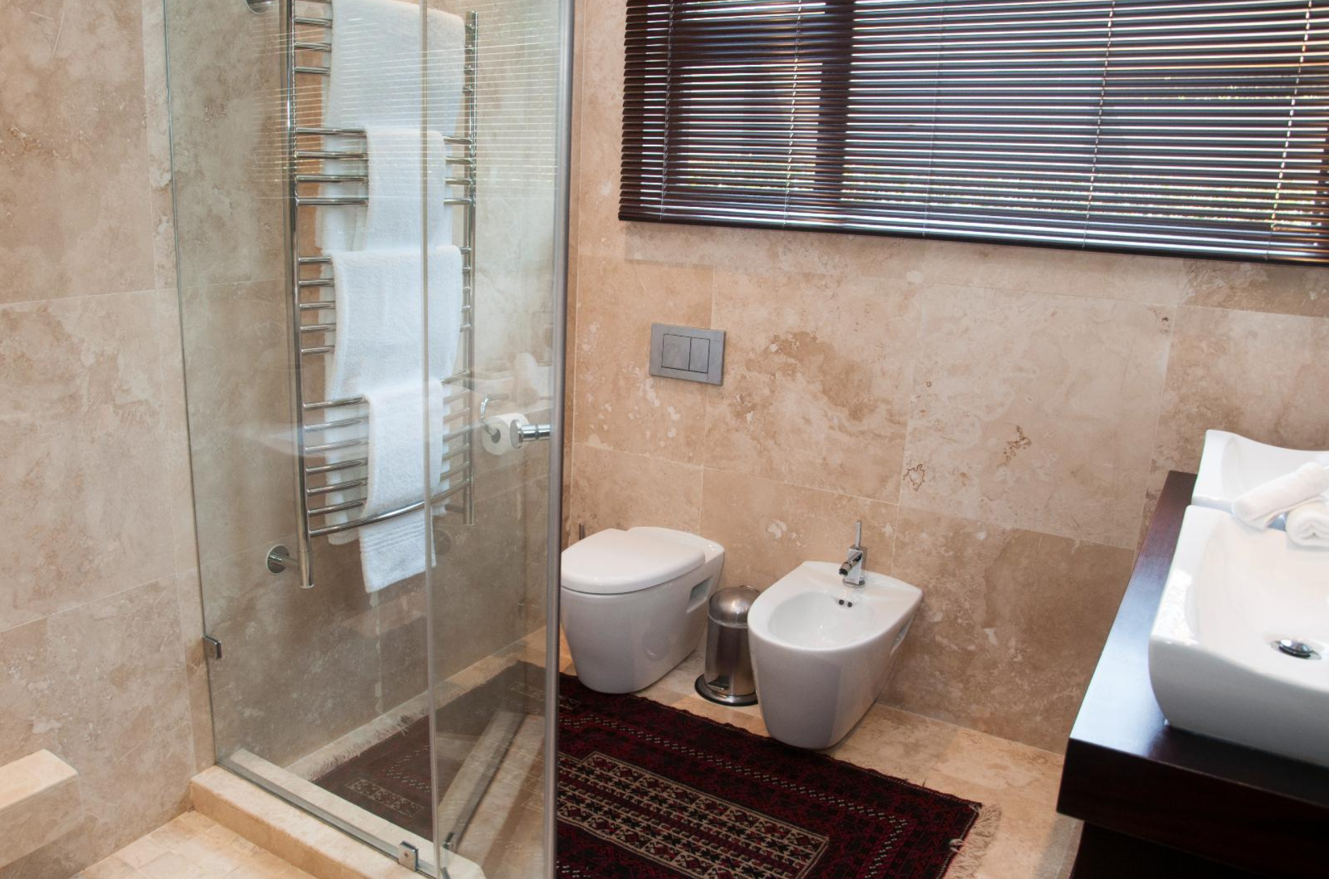 Bathroom with shower, toilet, bidet, sink, and wooden blinds. Beige stone walls and floor, towel warmer, red rug.