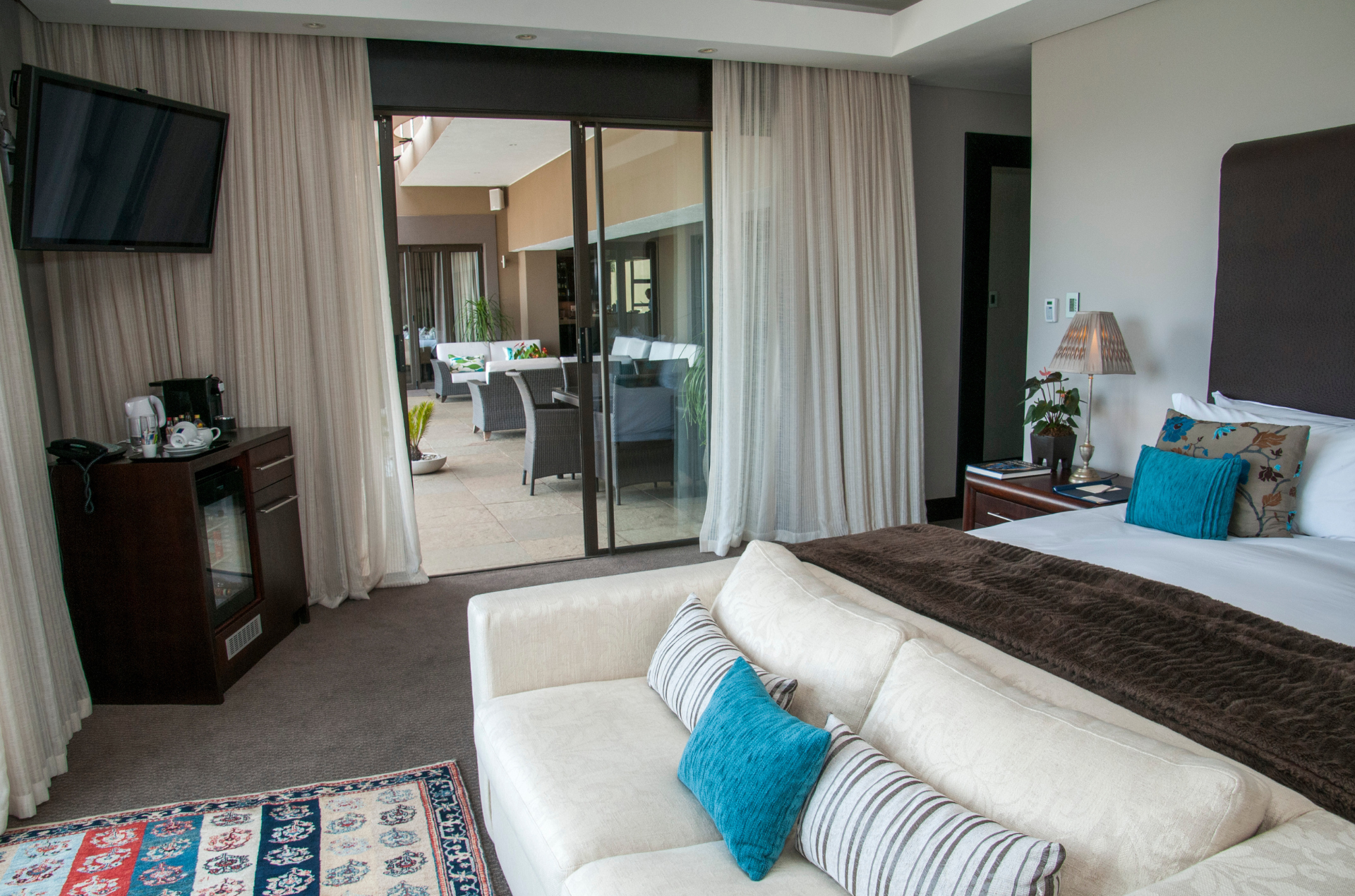 Hotel room with a bed, white couch, and sliding glass doors leading to a patio with outdoor seating.
