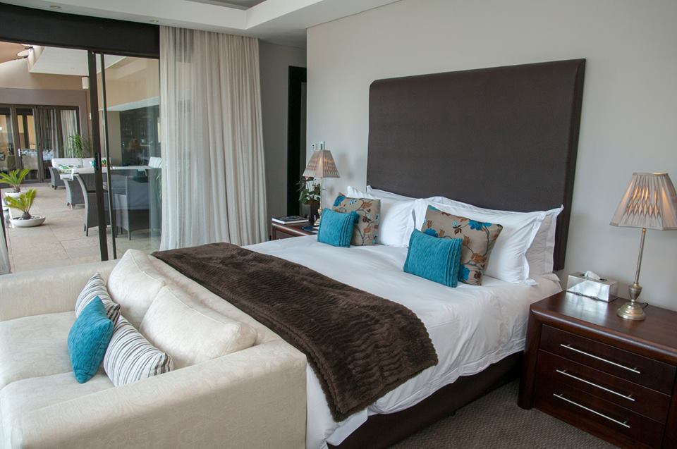 Bedroom with king-sized bed, white linens, brown headboard and throw, teal pillows, bedside tables, and a sliding door.