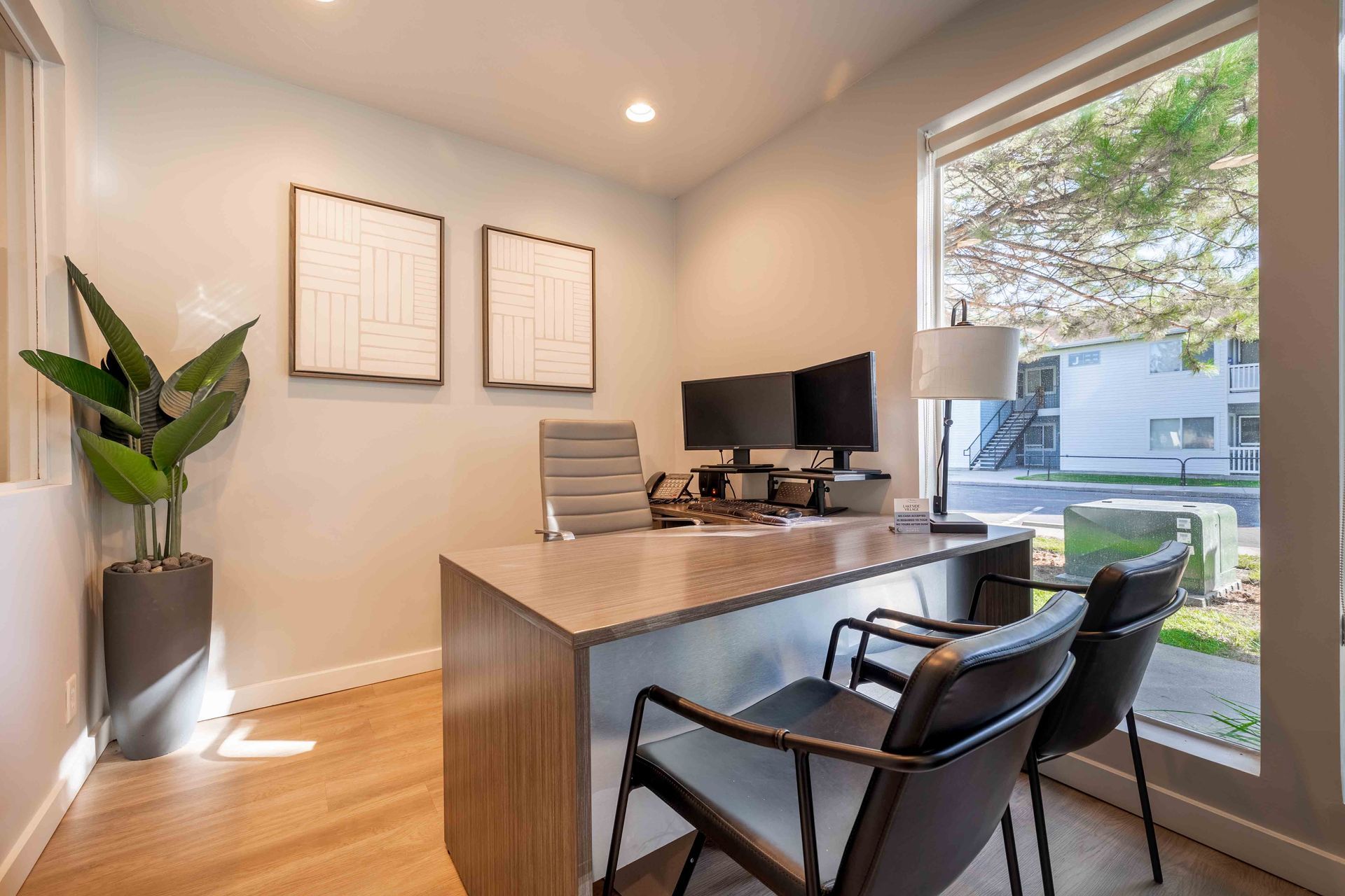 Office with desk, two monitors, and chairs. Large window with outdoor view, plant, and artwork.