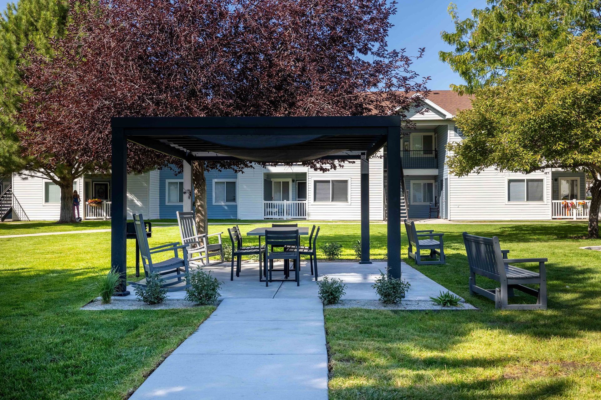 Outdoor seating area with pergola, table, chairs, and landscaping in front of a multi-story apartment building.