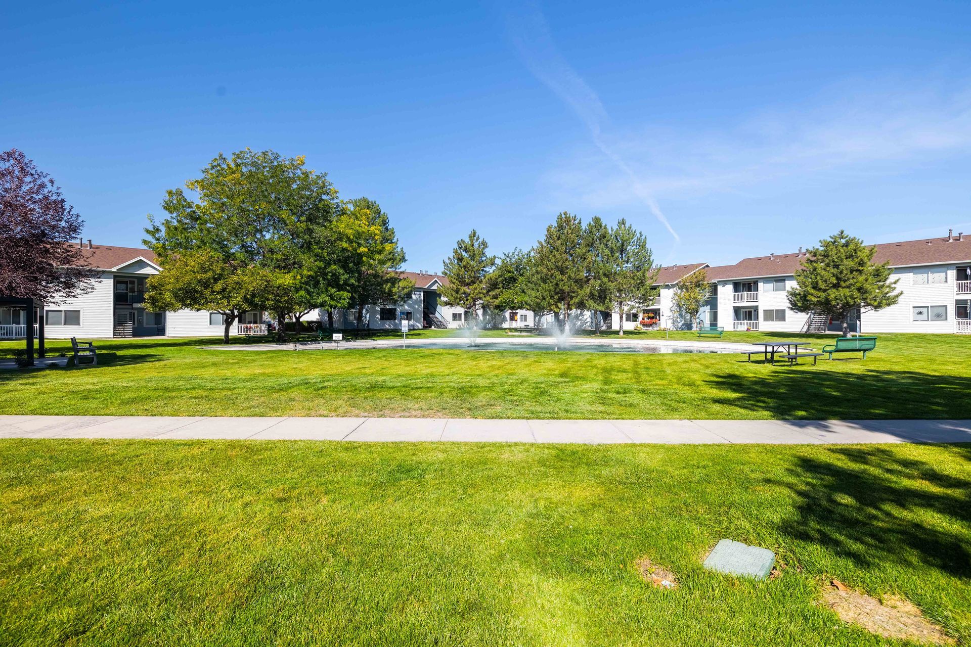 Green grassy area with fountain, trees, and two-story apartment buildings under a blue sky.