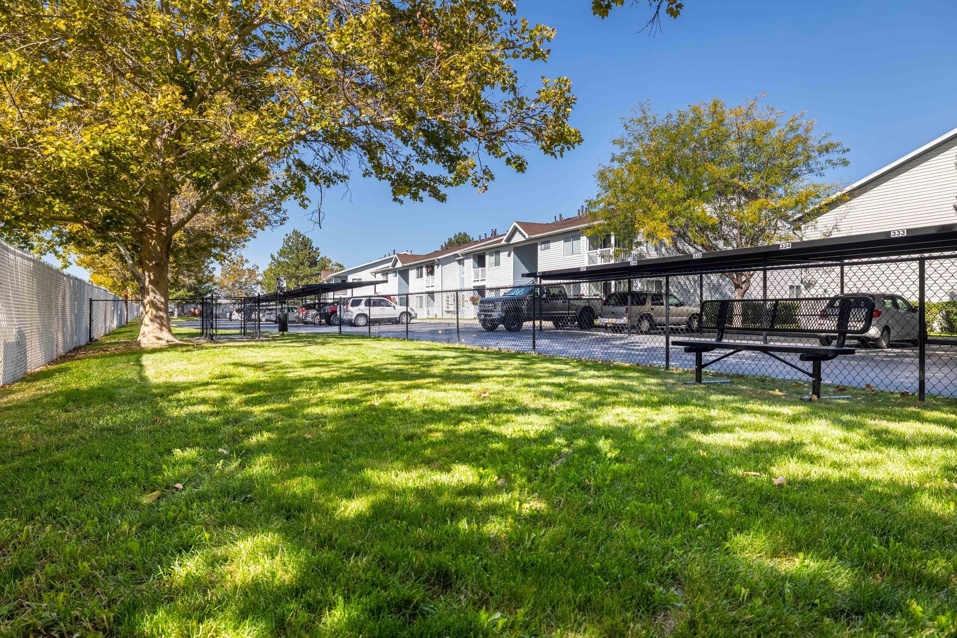 Lawn with covered parking spaces, cars parked, and apartment buildings in the background on a sunny day.