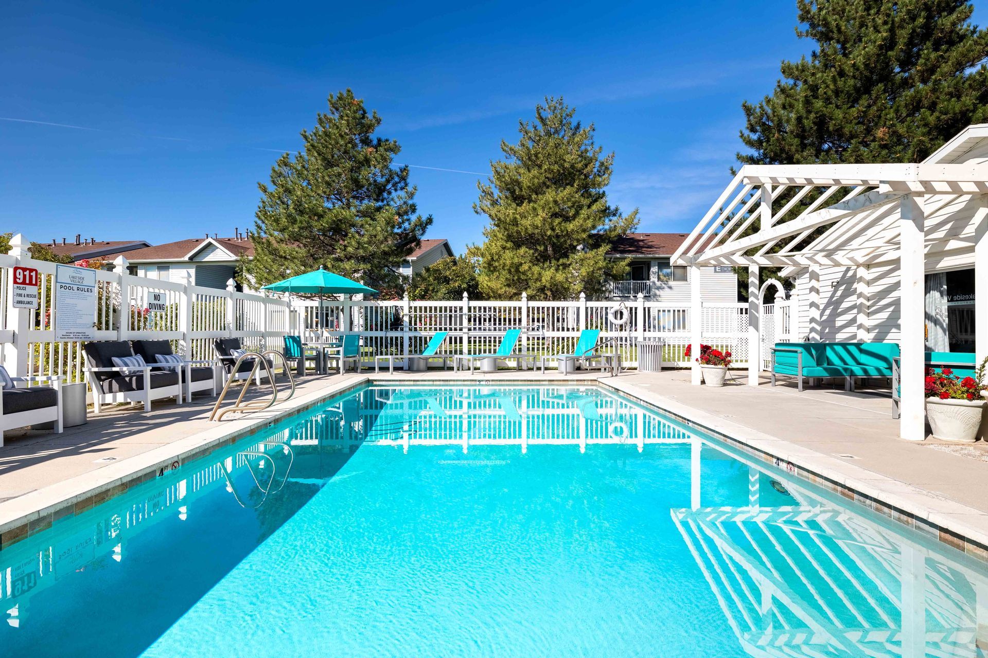 Swimming pool with turquoise water and white fence, chairs, and pergola under a blue sky.