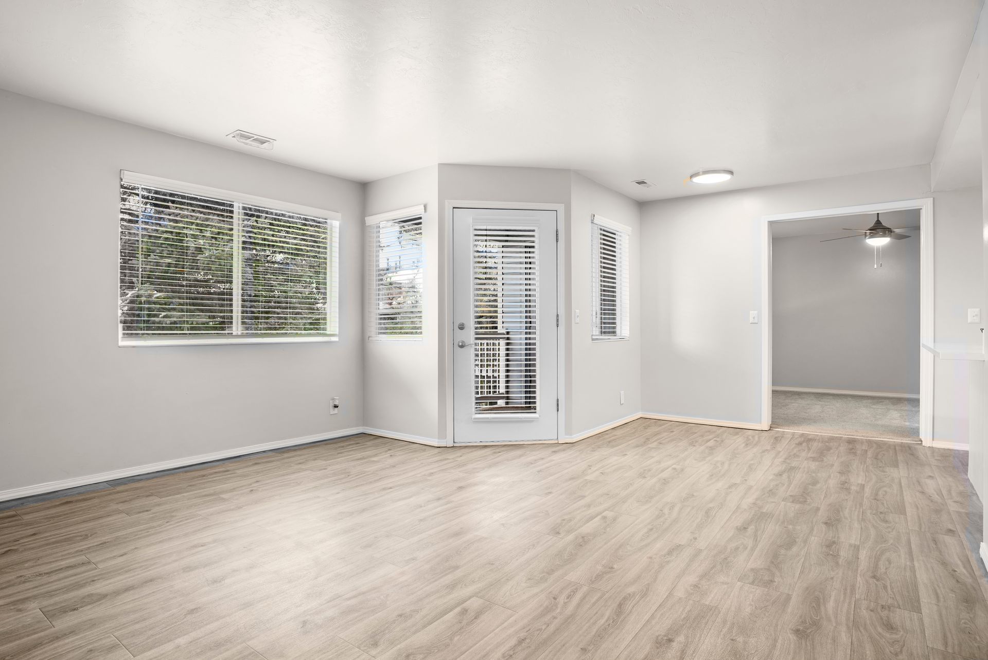 Empty, light-filled living room with light wood floors, large windows, and a doorway to another room.
