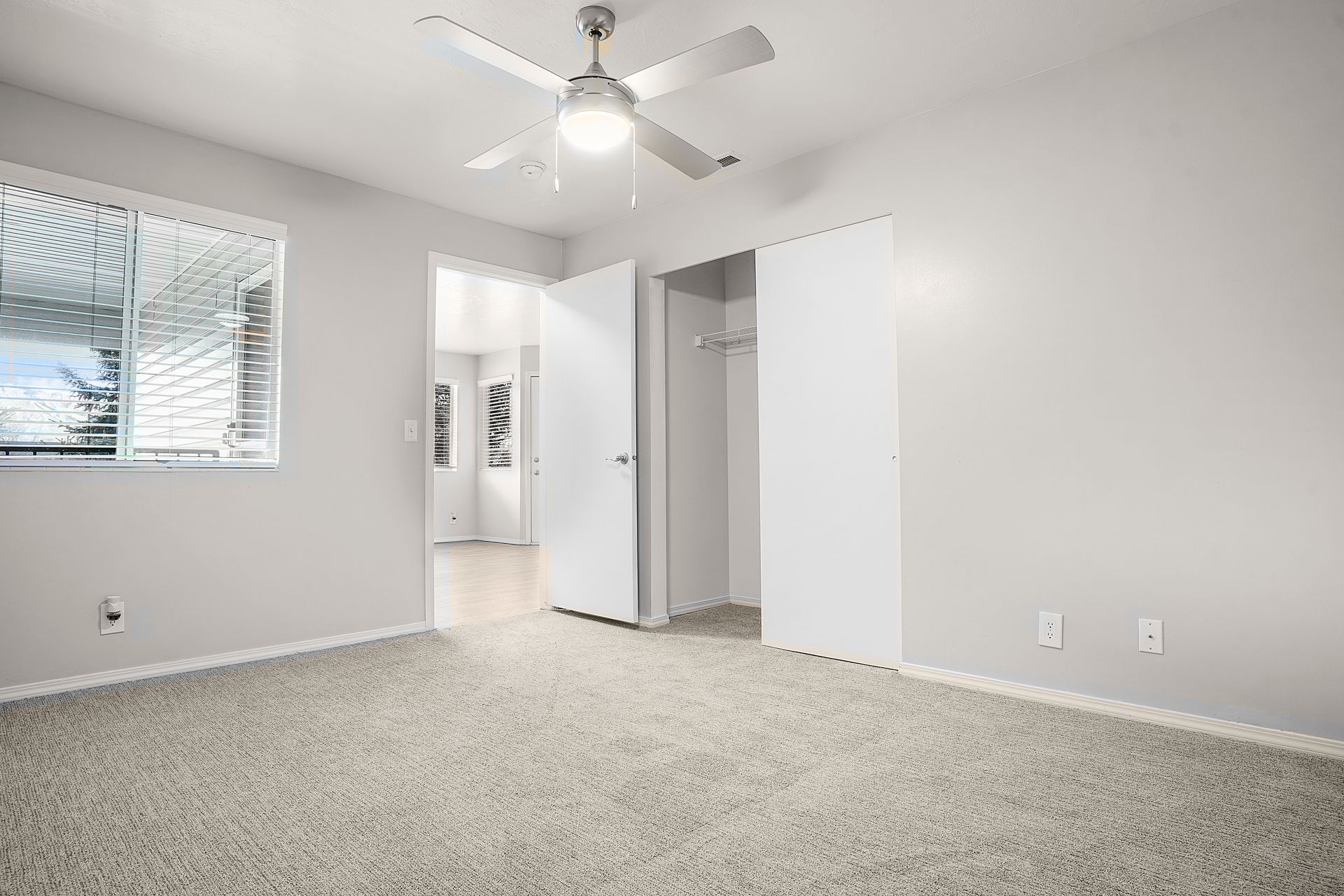 Empty bedroom with light grey walls, carpet, window, ceiling fan, and open closet.