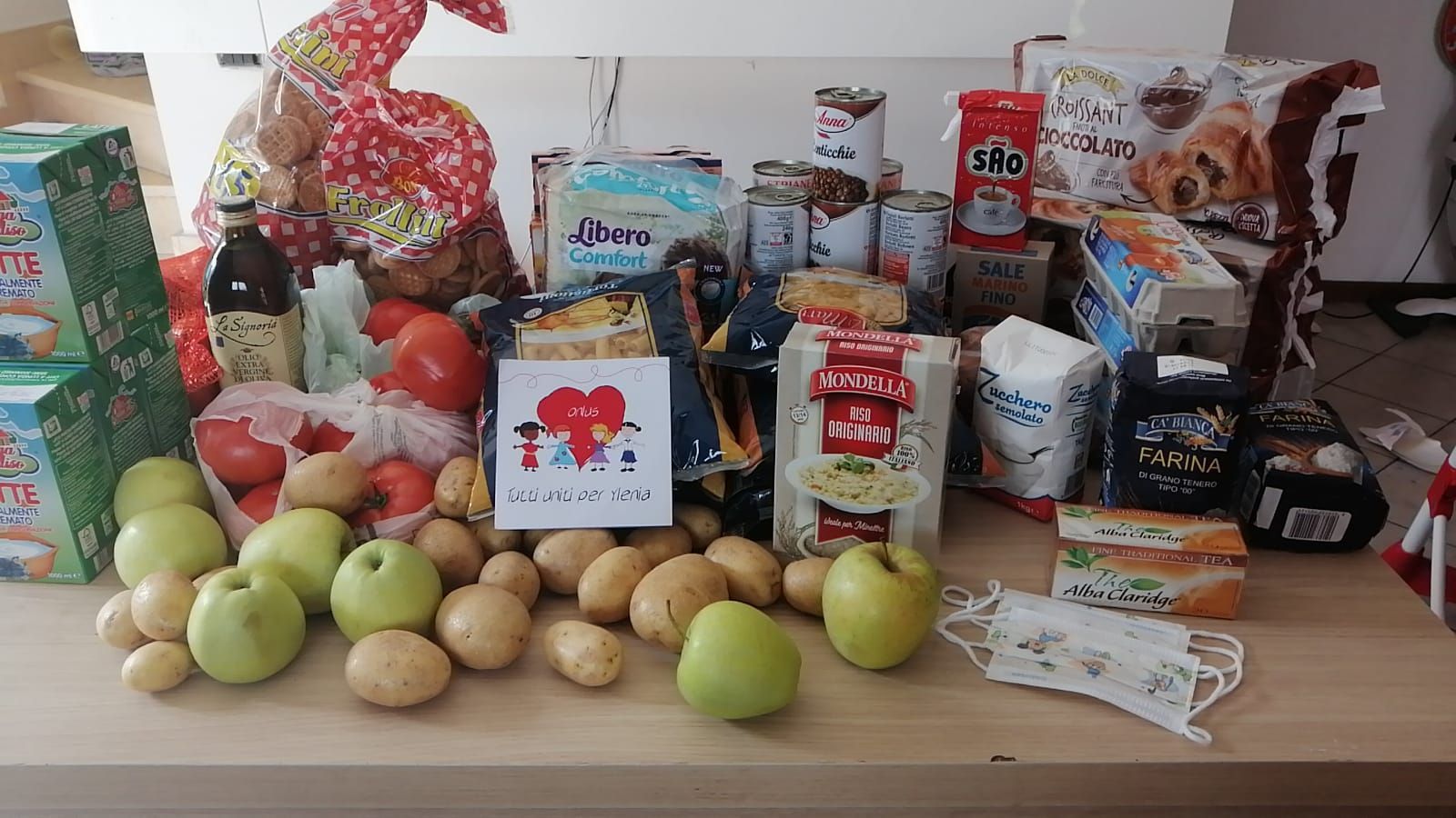 Una selezione di generi alimentari, tra cui prodotti freschi come mele e patate, conserve e un biglietto con l'illustrazione di un cuore.