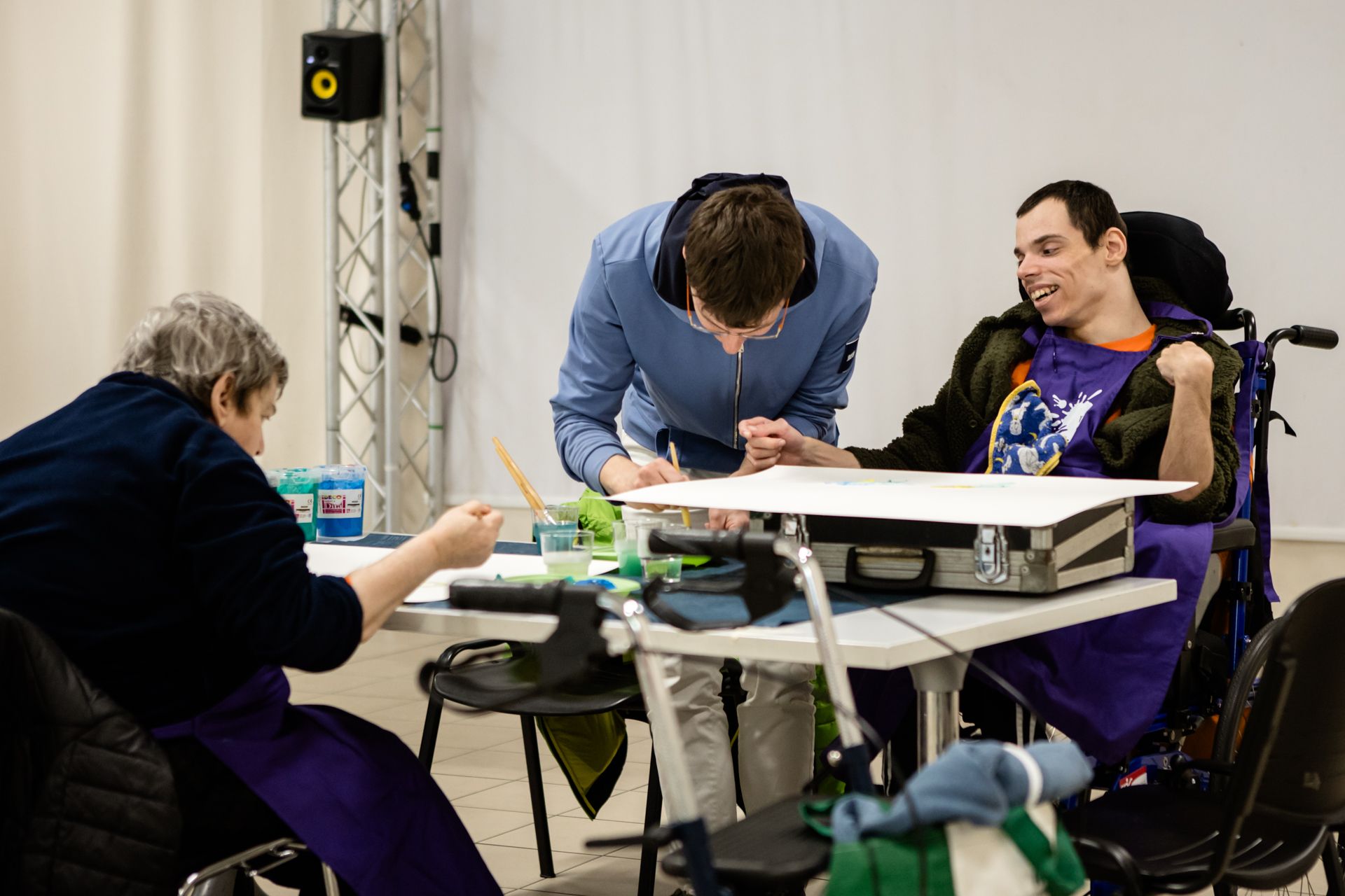 Tre persone siedono attorno a un tavolo bianco e creano opere d'arte; una di queste, in sedia a rotelle, sorride mentre dipinge su una grande tela.