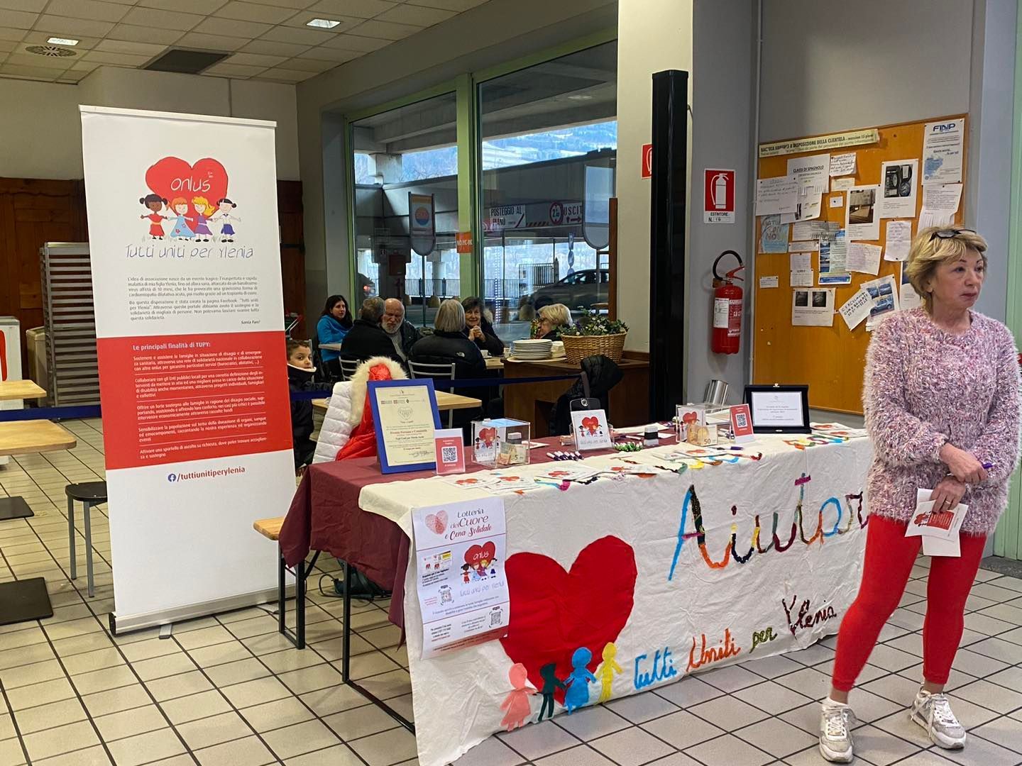 In una stanza luminosa, una donna è in piedi accanto a un tavolo informativo di beneficenza con uno striscione a forma di cuore con la scritta "Aiutar".