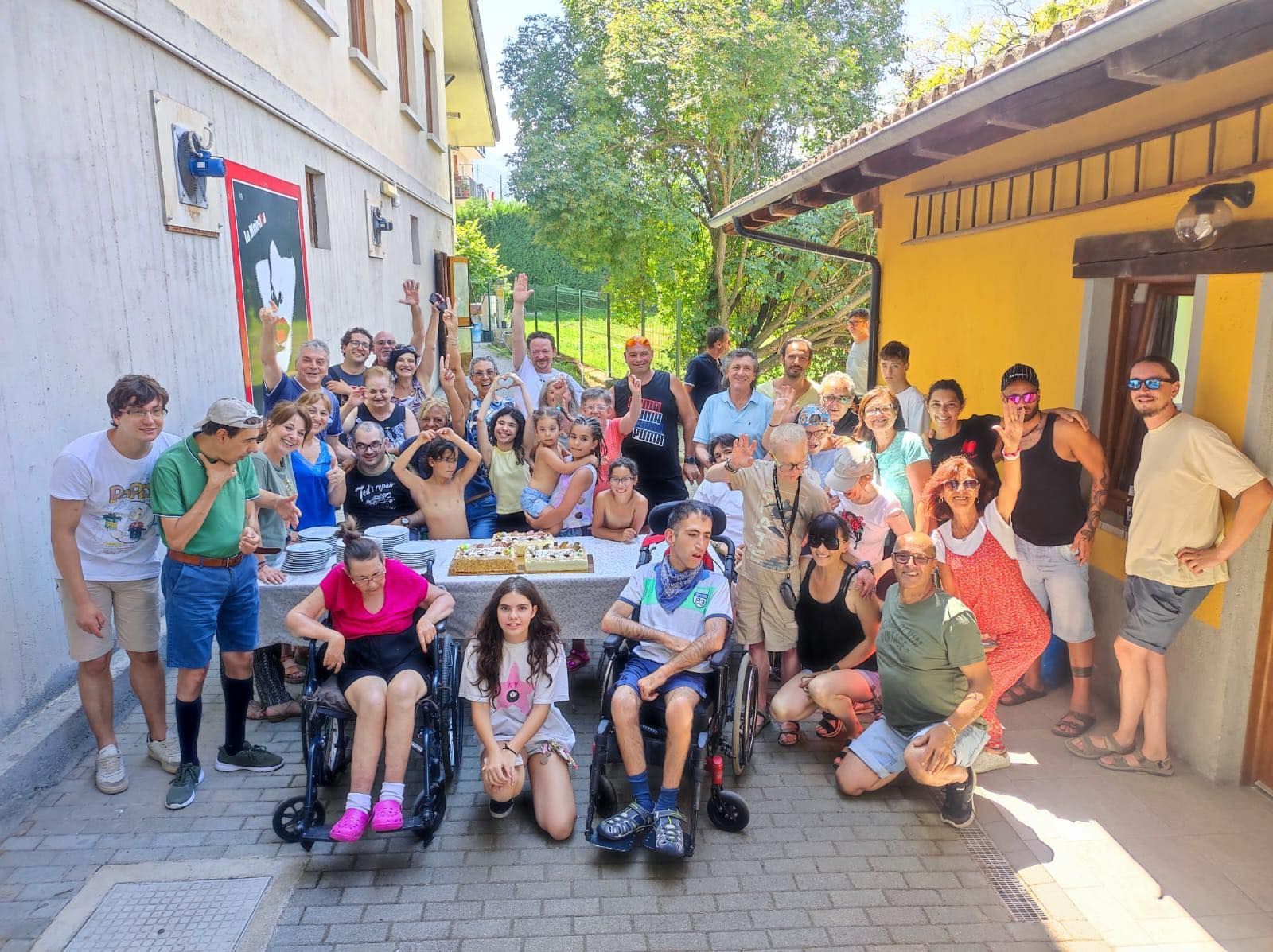 Un folto gruppo di persone posa per una foto all'aperto, davanti a edifici gialli e bianchi, vicino a un tavolo con una torta.