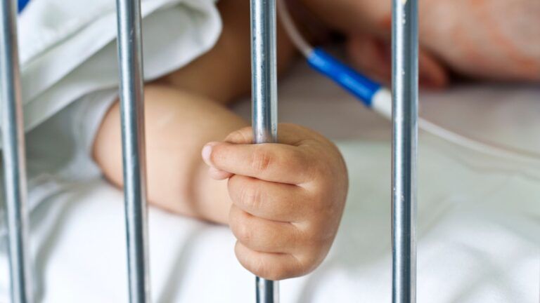 Una piccola mano afferra le sbarre verticali di una culla d'ospedale, con tubi medicali visibili sullo sfondo.