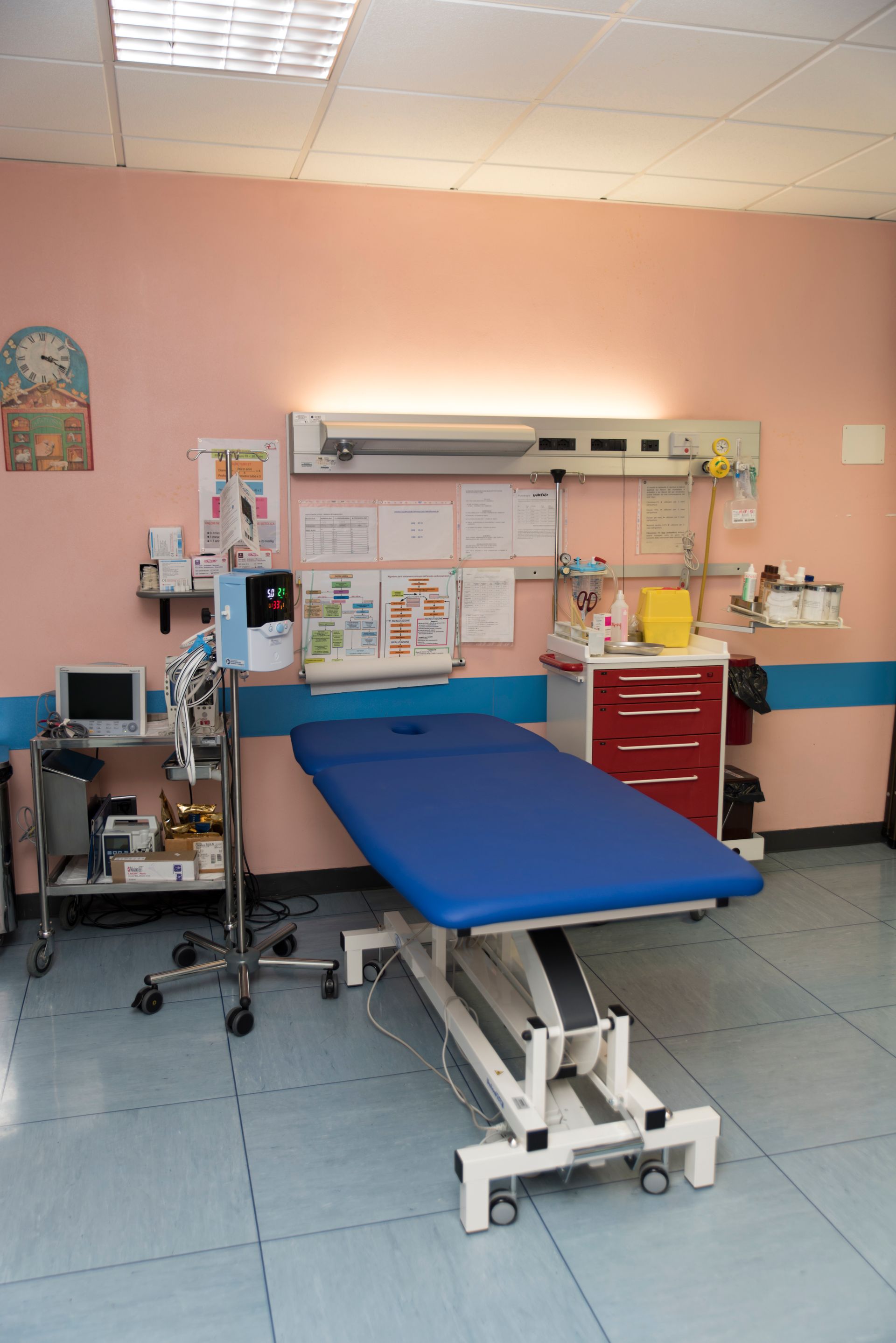 Una sala per visite mediche con un lettino blu, attrezzature mediche, un carrello rosso per il deposito e poster su una parete rosa.