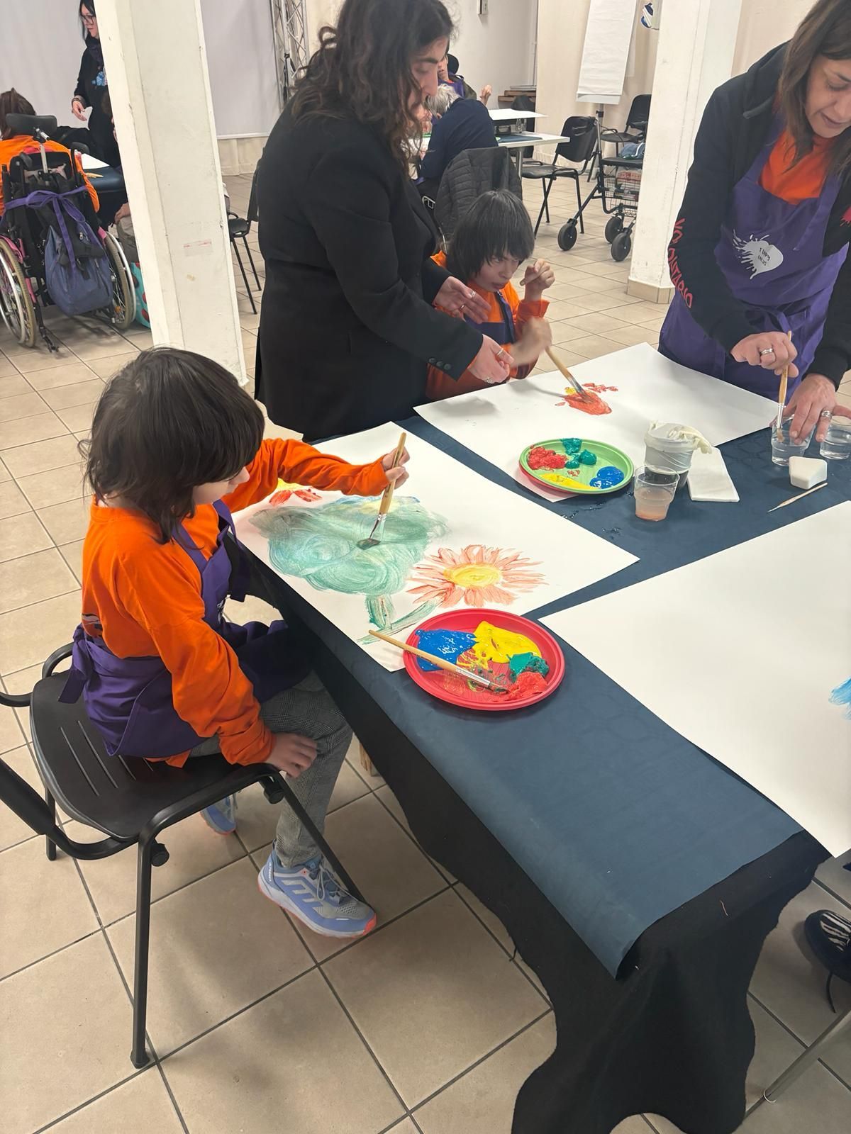 In una stanza ben illuminata, alcune persone con il grembiule dipingono ai tavoli; una persona è seduta su una sedia a rotelle mentre le altre sono in piedi nelle vicinanze.