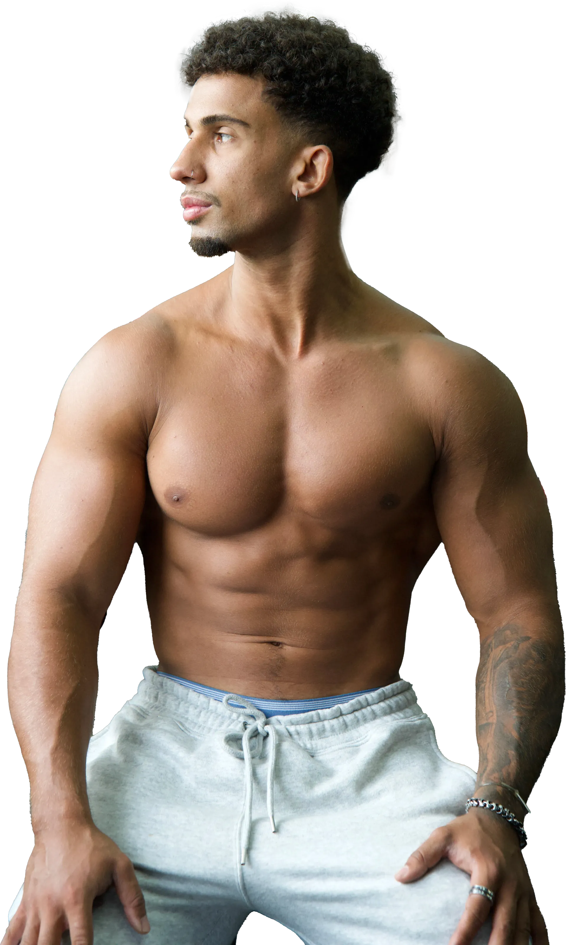 Muscular man with dark, curly hair, looking off to the side, bare-chested, wearing gray sweatpants, indoors. JUNIOR WORKOUT