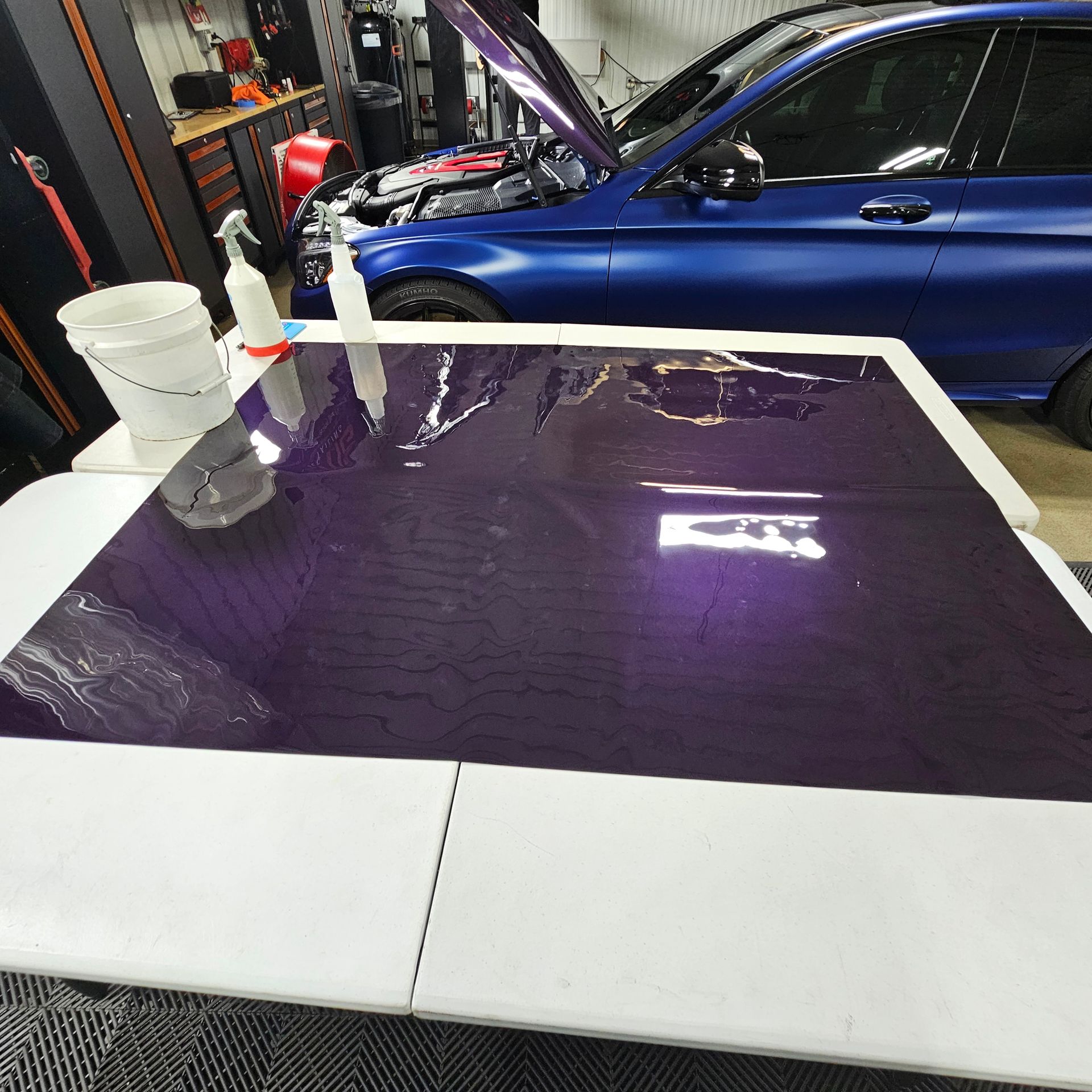 A sheet of purple vinyl on a white table, next to a blue car with its hood open, and a bucket.