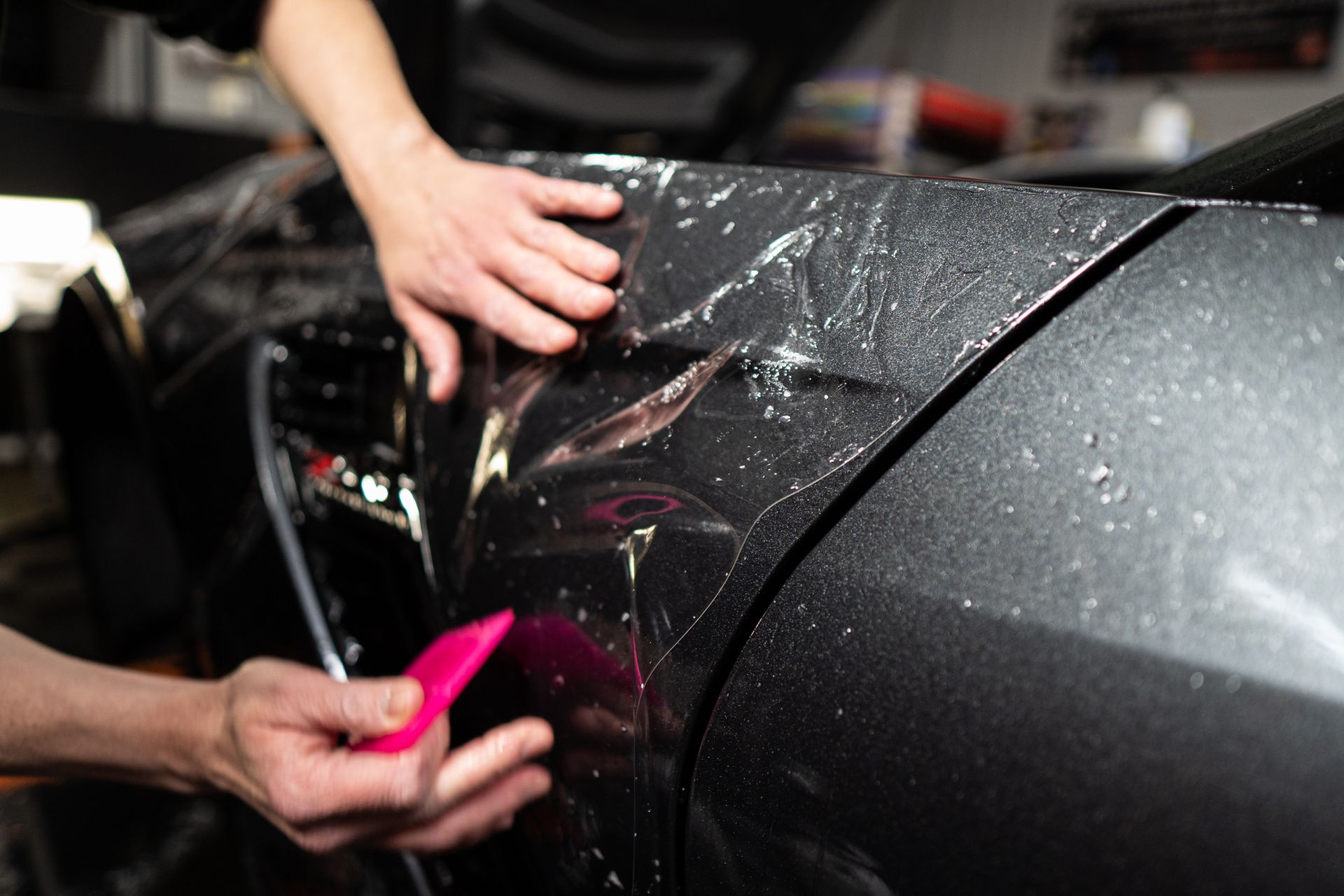 Hands applying protective film to a car's gray fender with a pink squeegee.