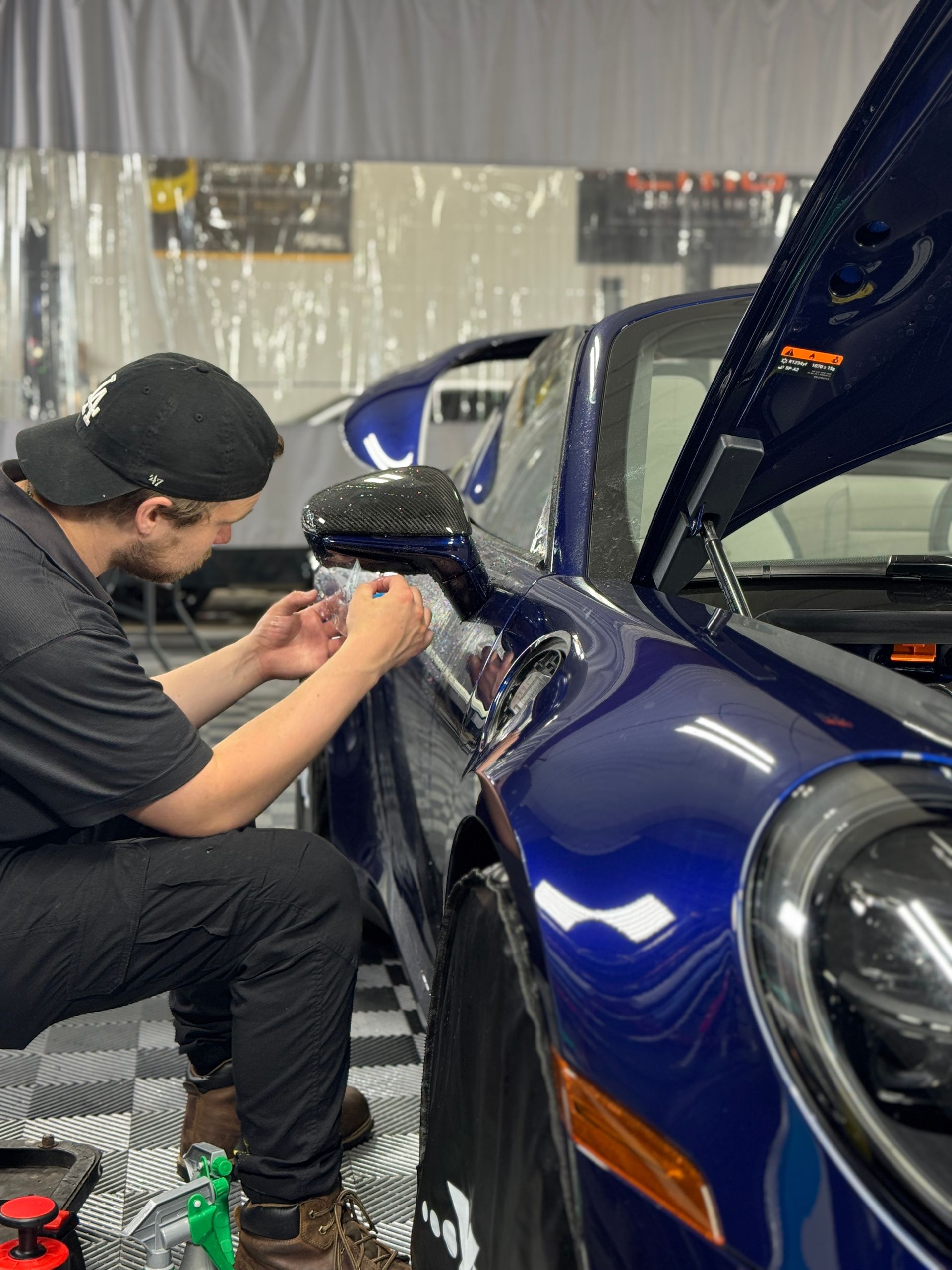 A person installing something on a blue sports car in a garage. The hood is up.